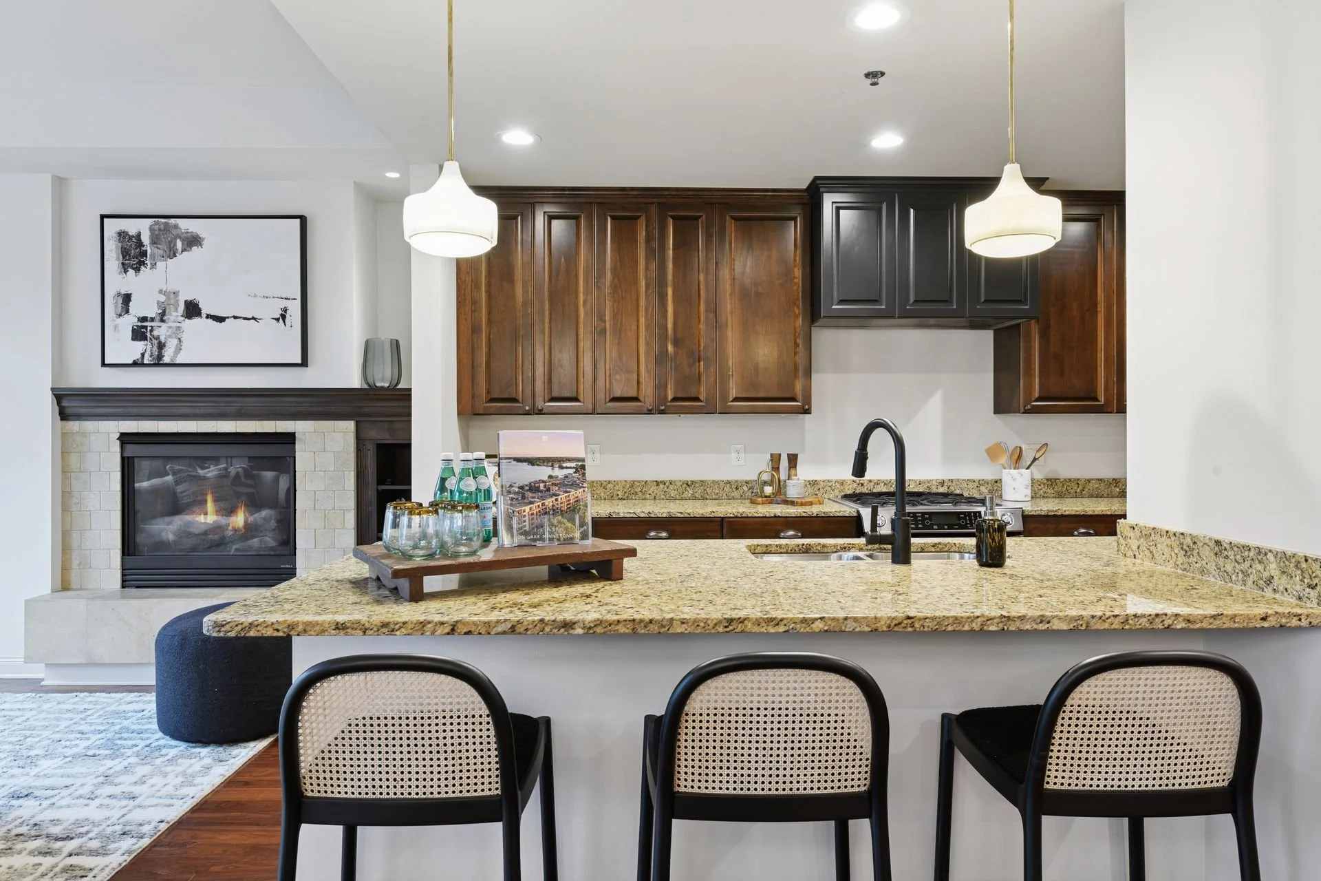 Modern kitchen with granite countertops, dark wooden and black cabinetry, a black faucet, and two pendant lights, with a view of a living room with a fireplace.