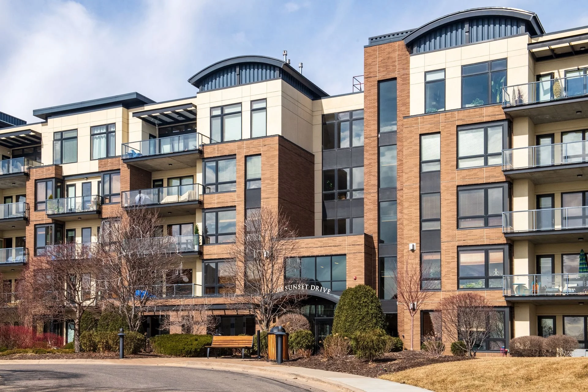 Multi-story apartment building with balconies, brick and glass facade, and a landscaped entrance with trees and bushes.