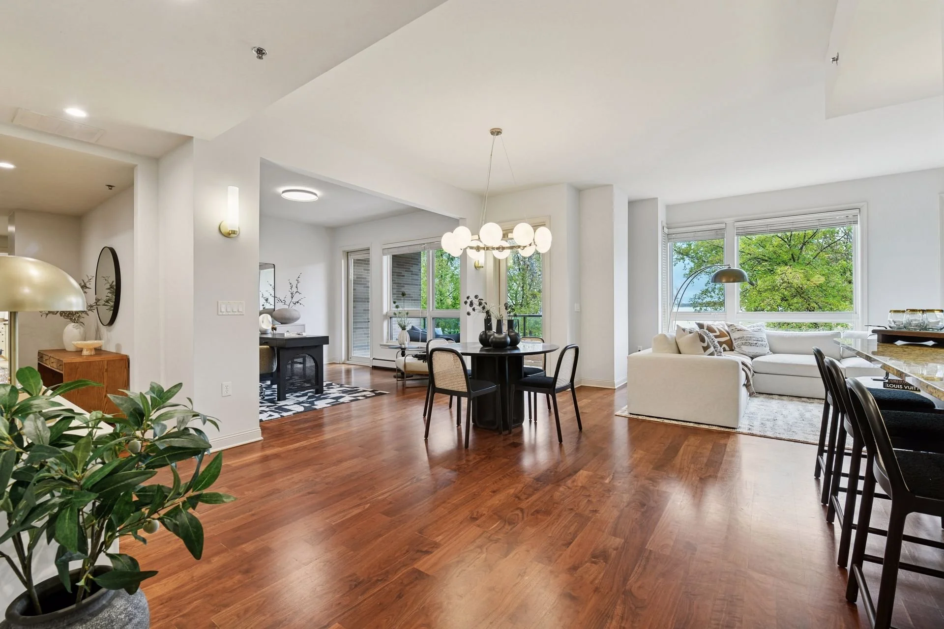 Bright open-plan living and dining area with hardwood floors, large windows showing green trees, and stylish lighting fixtures.
