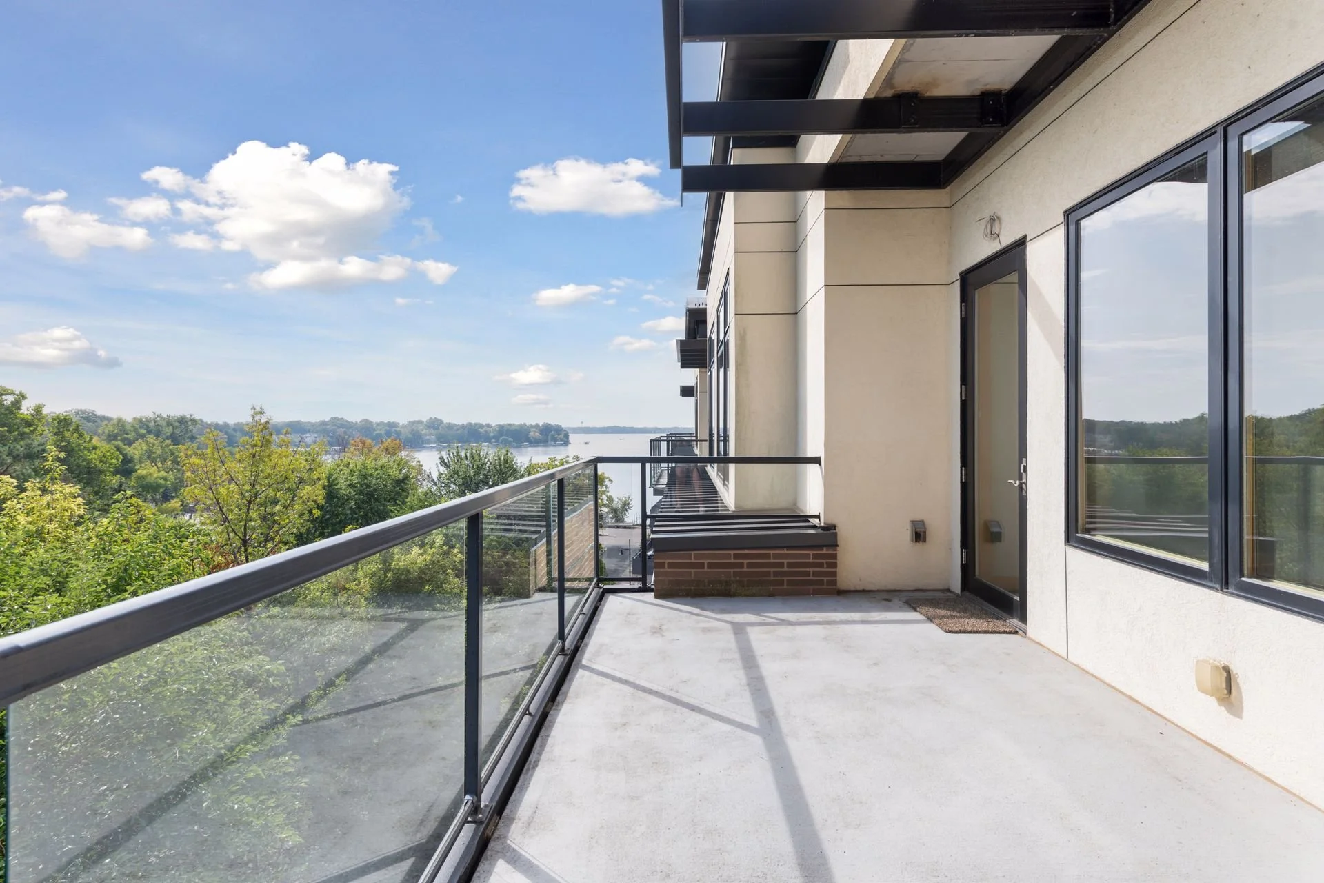 A balcony with a glass railing overlooking a scenic view of trees and a river under a partly cloudy sky.