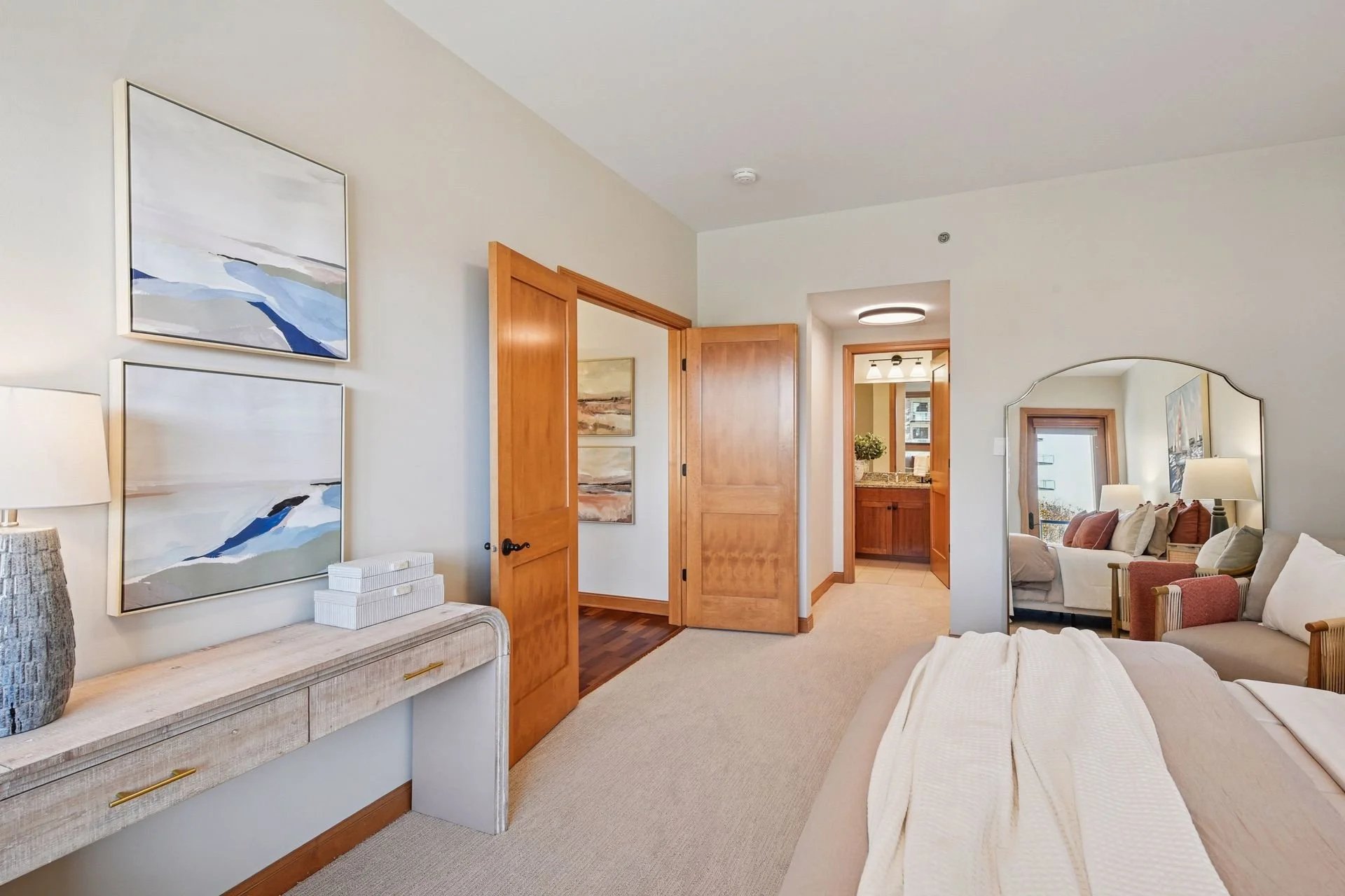 A bedroom with light-colored walls. Wooden doors open to a bathroom with vanity and a window showing outdoor scenery.