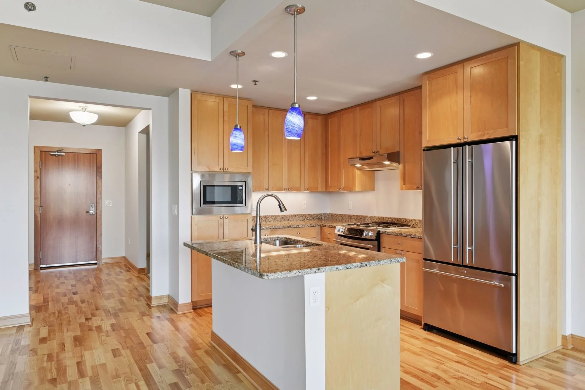 Modern kitchen with wood cabinetry, stainless steel refrigerator, microwave, granite countertops, and hardwood flooring, illuminated by pendant and recessed lighting.