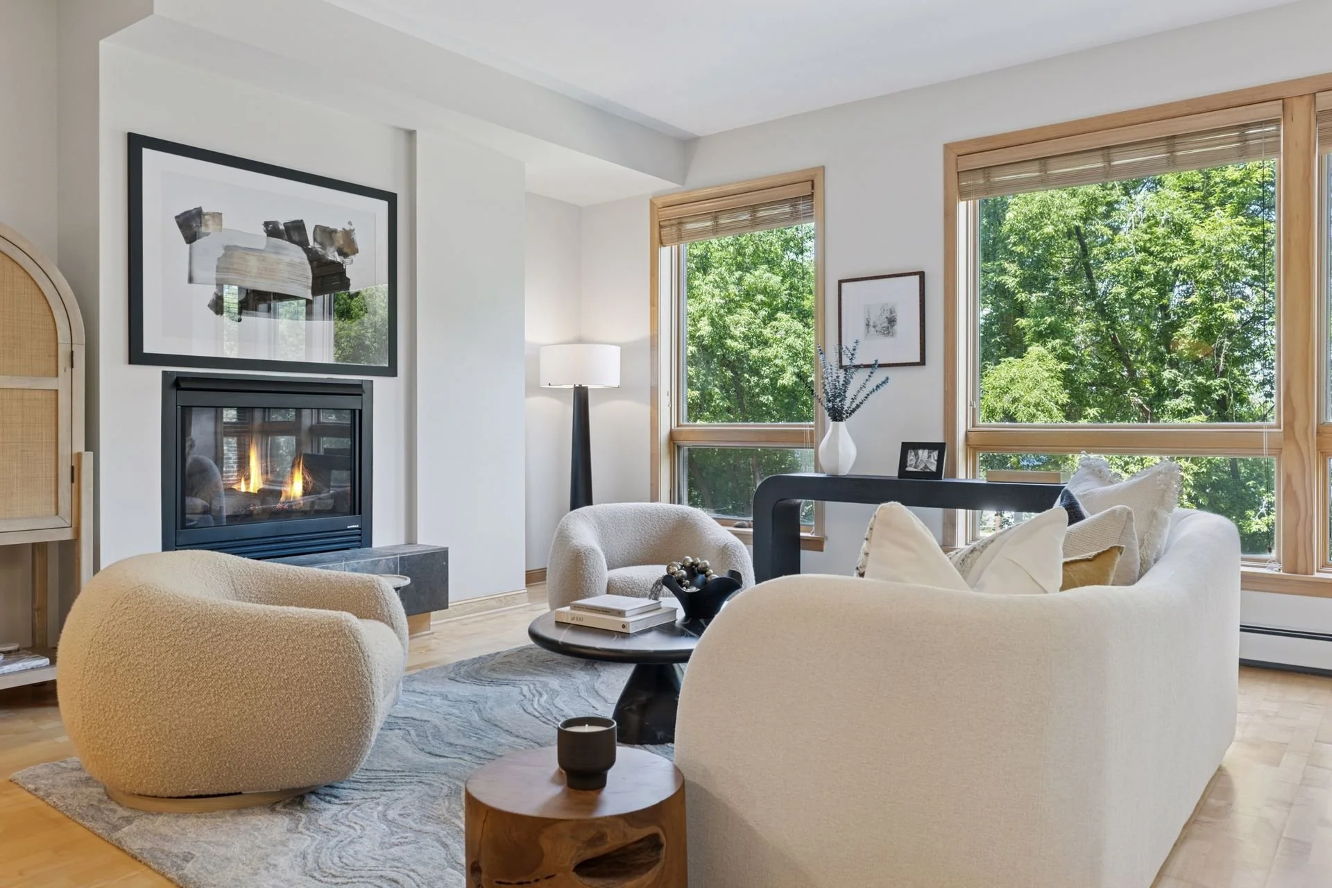 Living room with a fireplace, large windows, and a view of green trees outside.