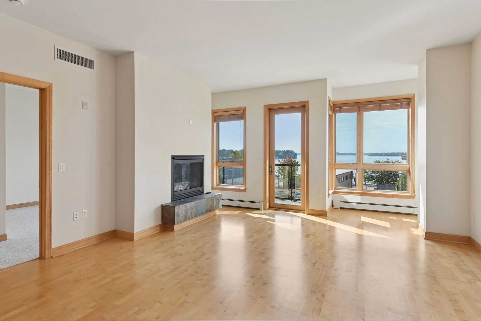 Empty living room with hardwood floors, large windows and a view of a lake, featuring a fireplace with a stone base and wooden trim around windows and door.