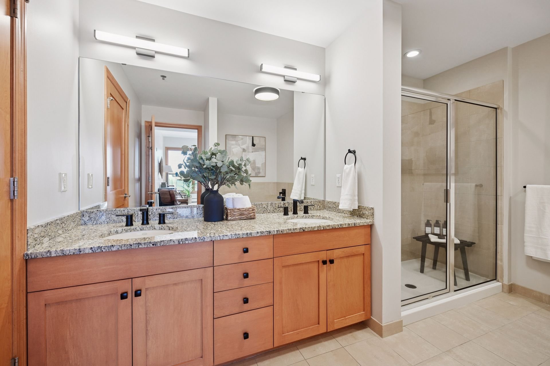 Bathroom with wooden vanity, granite countertop, large mirror, black fixtures, and a glass shower with a seat.
