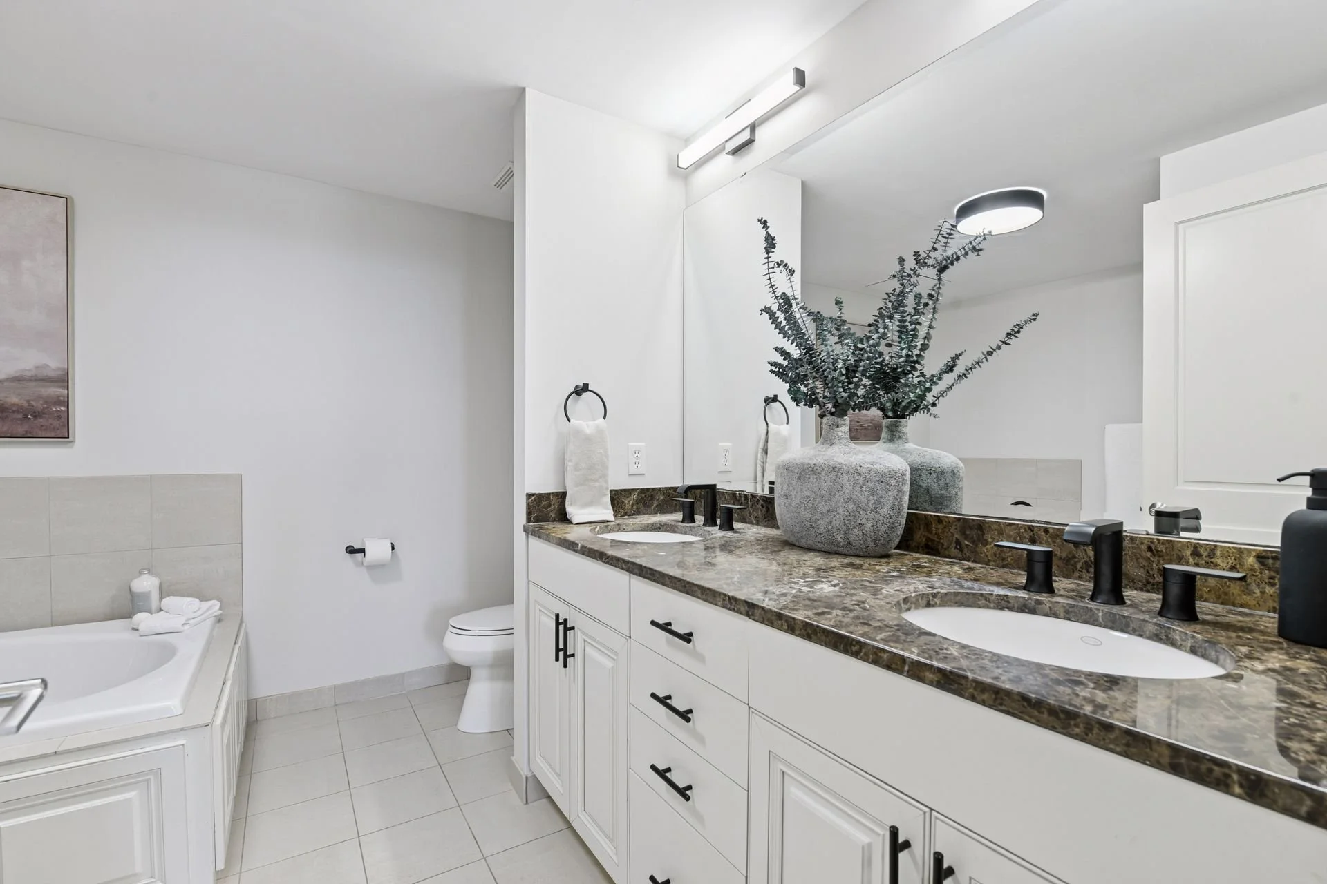 Modern bathroom with white cabinetry, a granite countertop, two sinks, black fixtures, a large mirror, a bathtub, and a toilet.