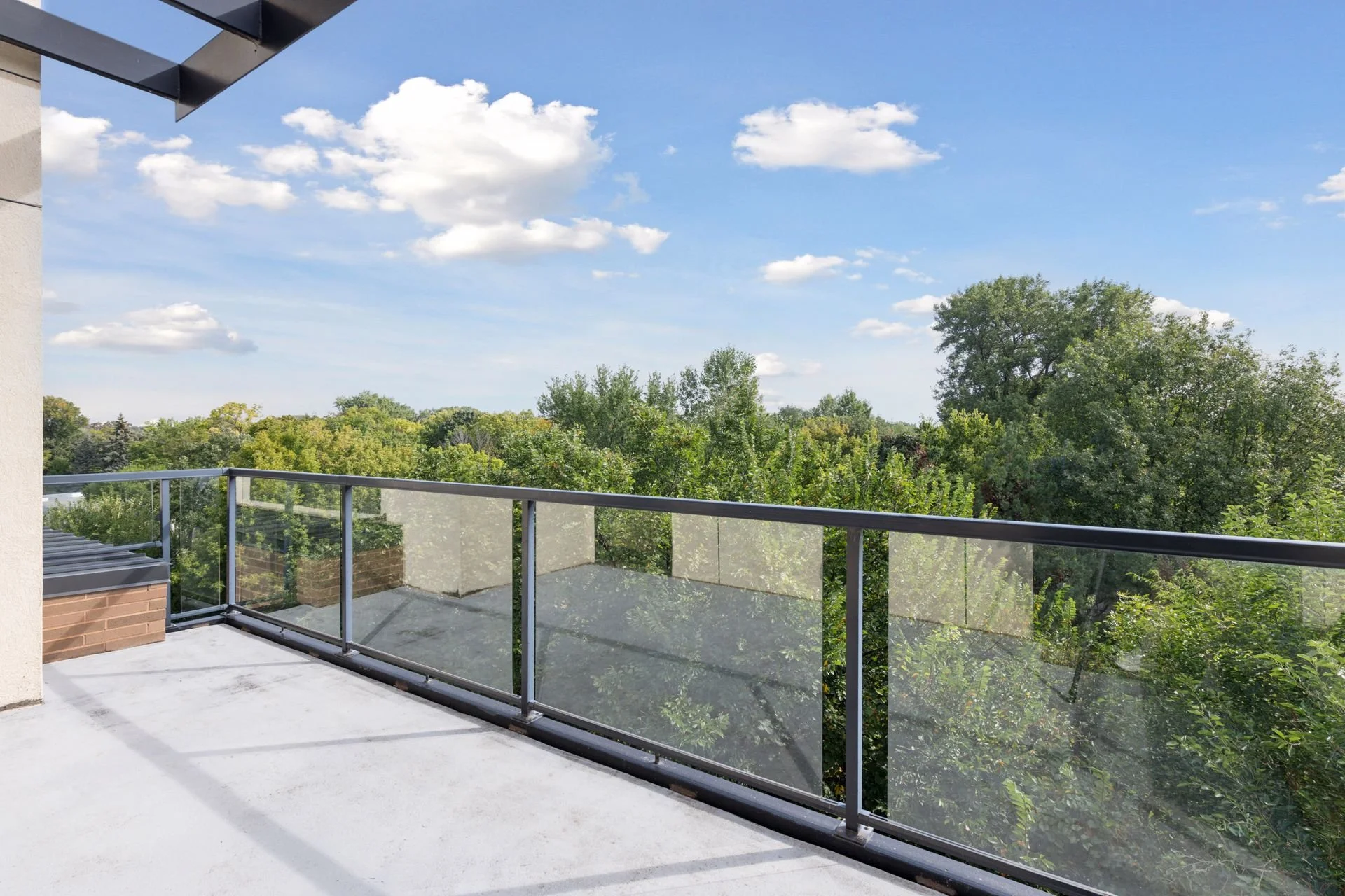 View from a balcony with a glass railing overlooking green trees and a blue sky with scattered clouds.