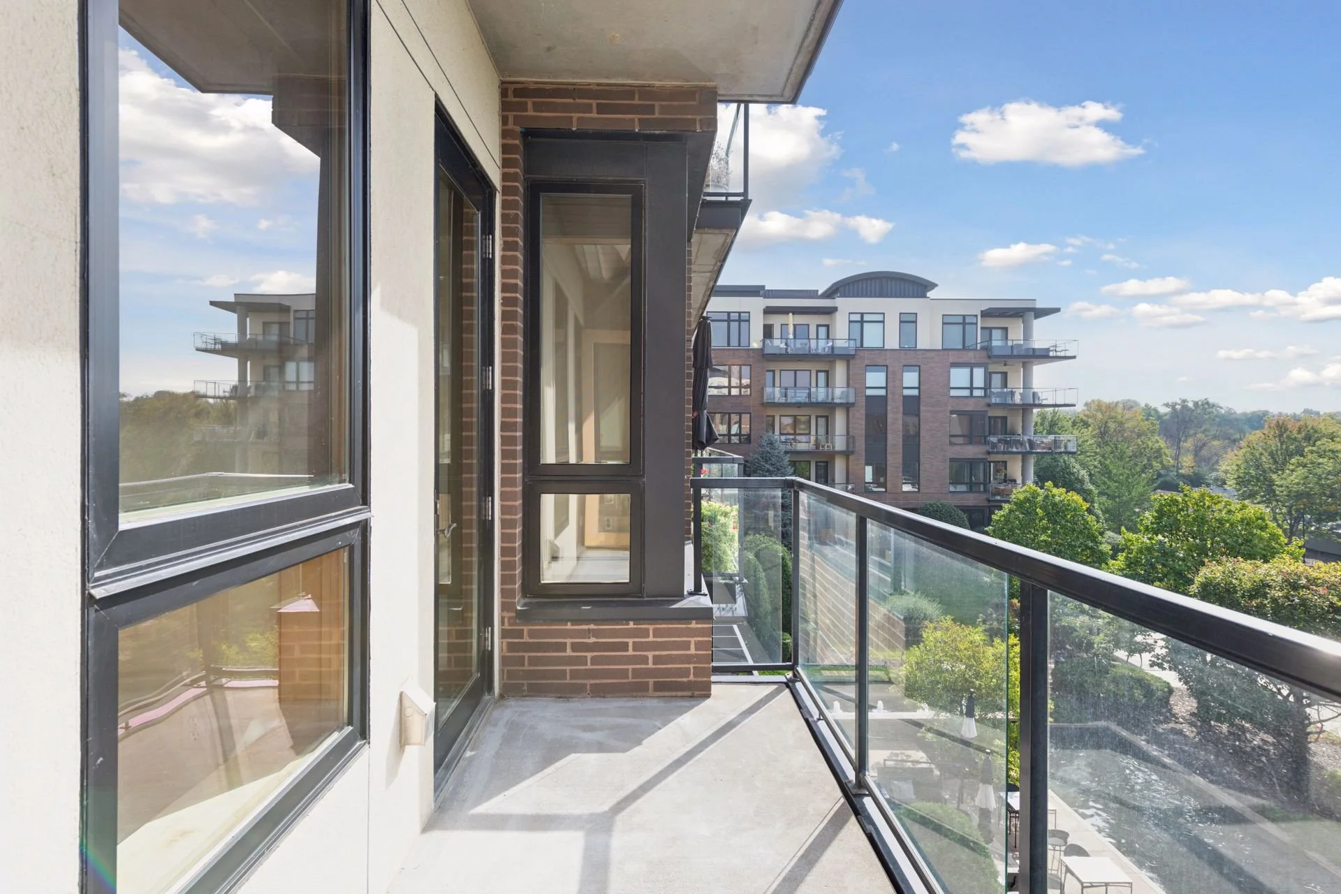 View from a balcony of a modern apartment building with glass railing, neighboring building, trees, and a partly cloudy sky.