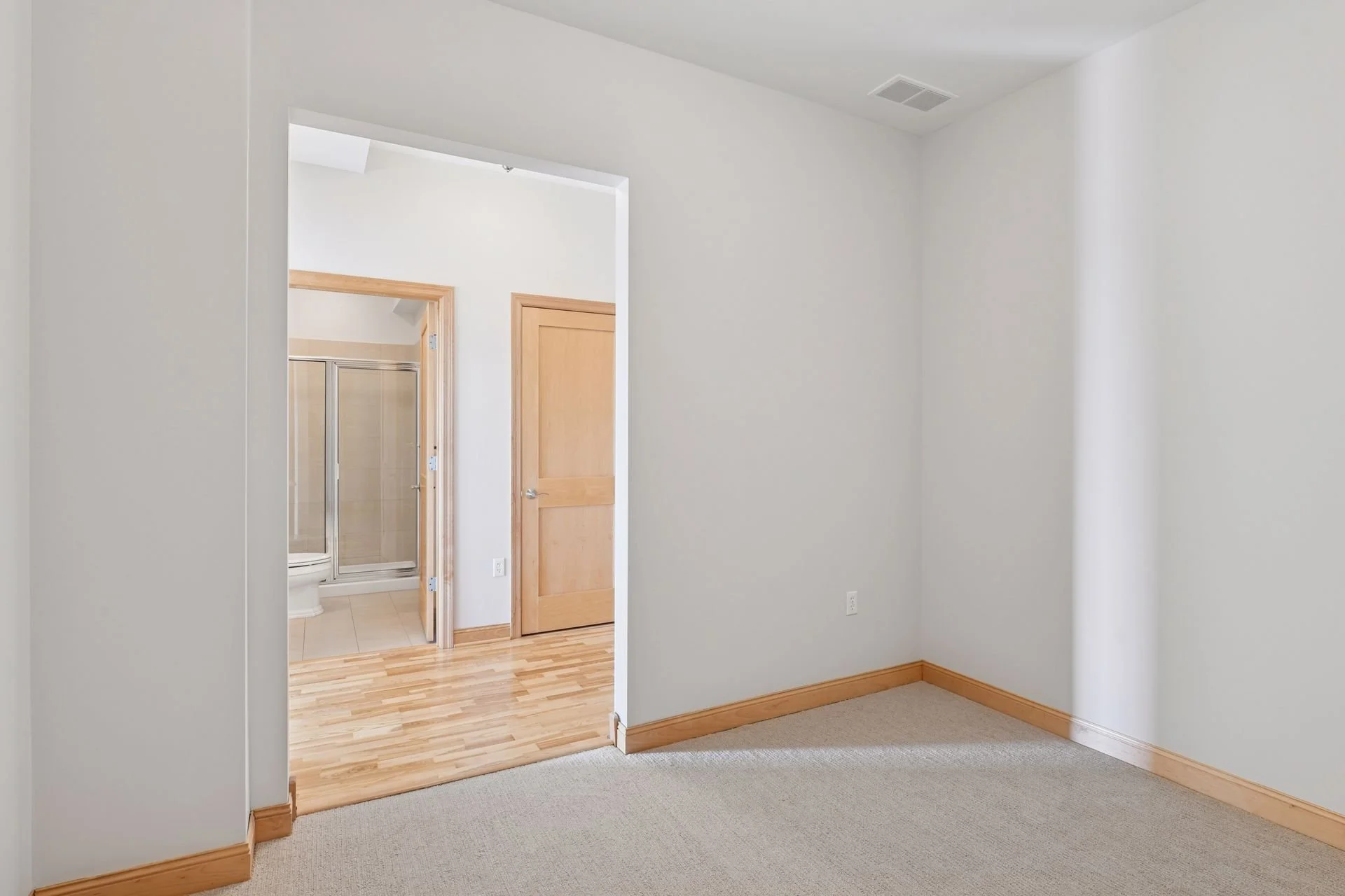 Empty room with beige carpet, white walls, wooden trim, and a doorway leading to a bathroom with shower stalls and a wooden door.