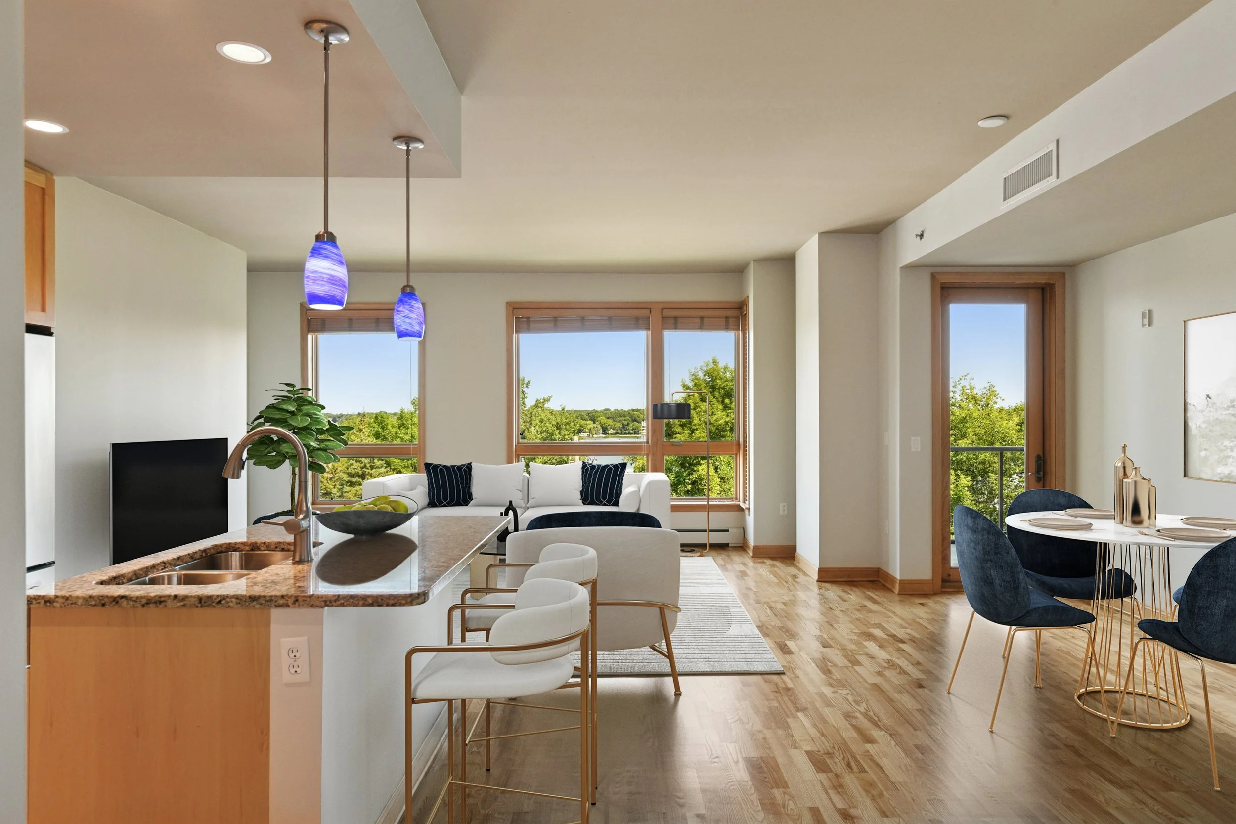 Open-concept living room and kitchen with hardwood floors and large windows showing green trees outside.