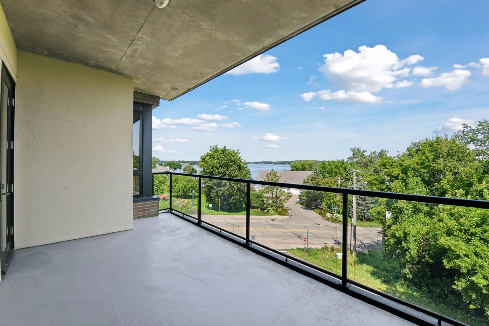 Balcony with black railing overlooking trees, a parking lot, and Lake Minnetonka in the distance under a blue sky with scattered clouds.