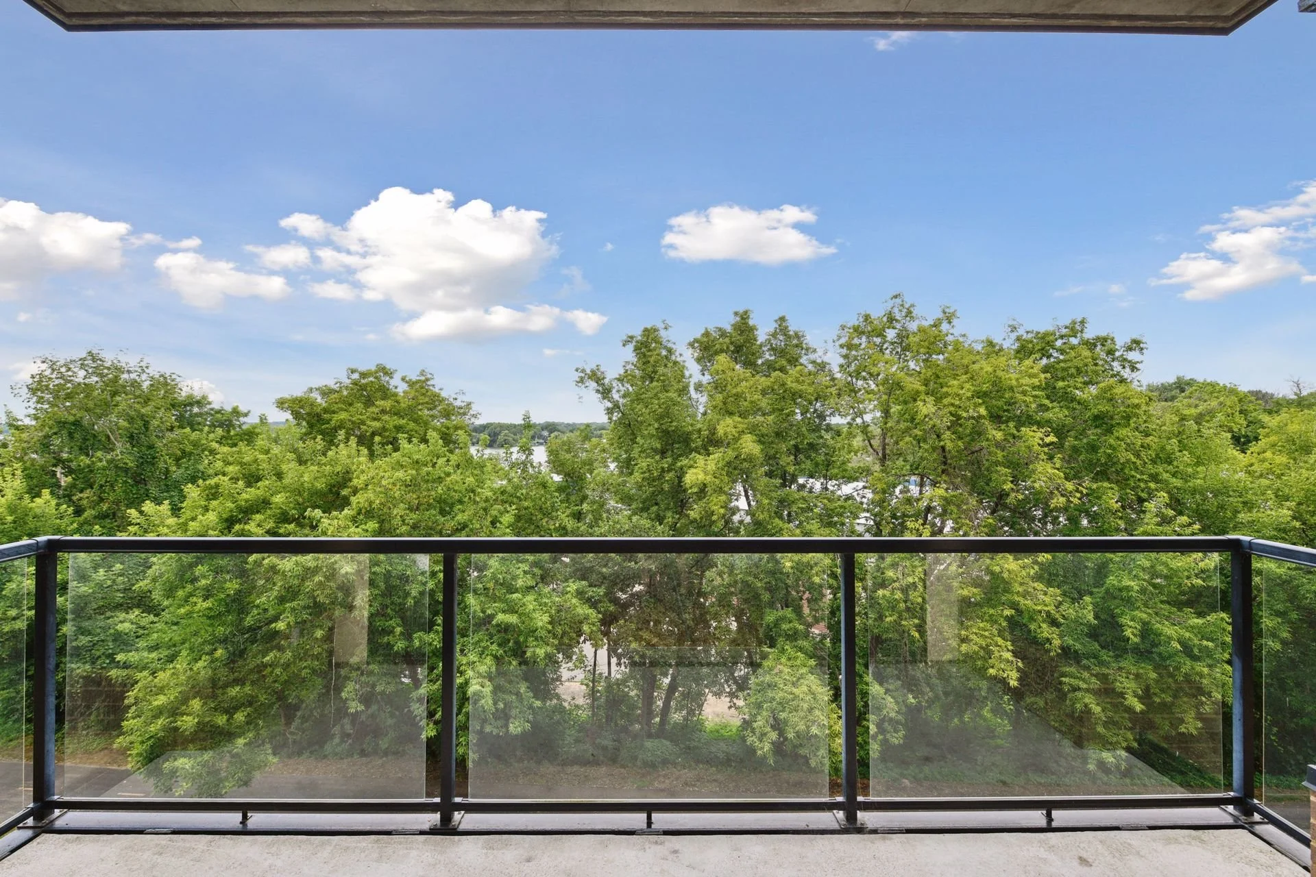 Balcony view overlooking lush green trees and a bright blue sky with scattered white clouds.