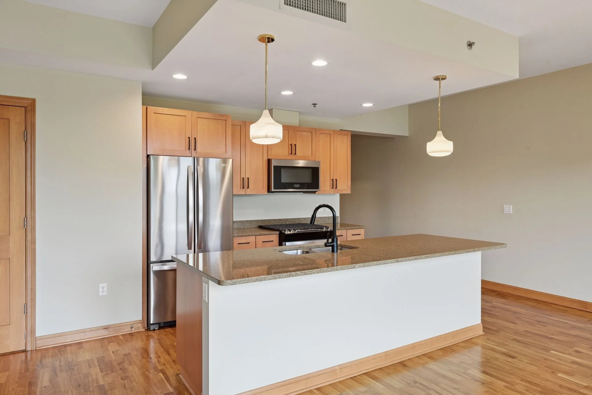 Modern kitchen with wooden cabinets, stainless steel refrigerator, microwave, granite countertops, and a kitchen island with a black faucet. The kitchen has hardwood floors and white walls, with pendant lights hanging from the ceiling.