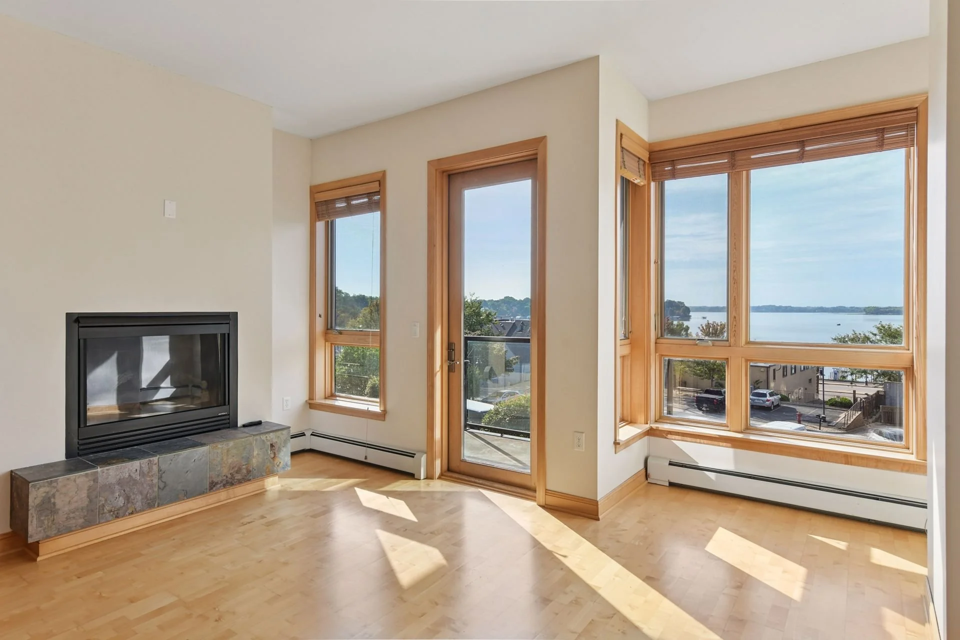 Bright living room with large windows overlooking water and trees, wooden trim, and a fireplace with a stone mantel.