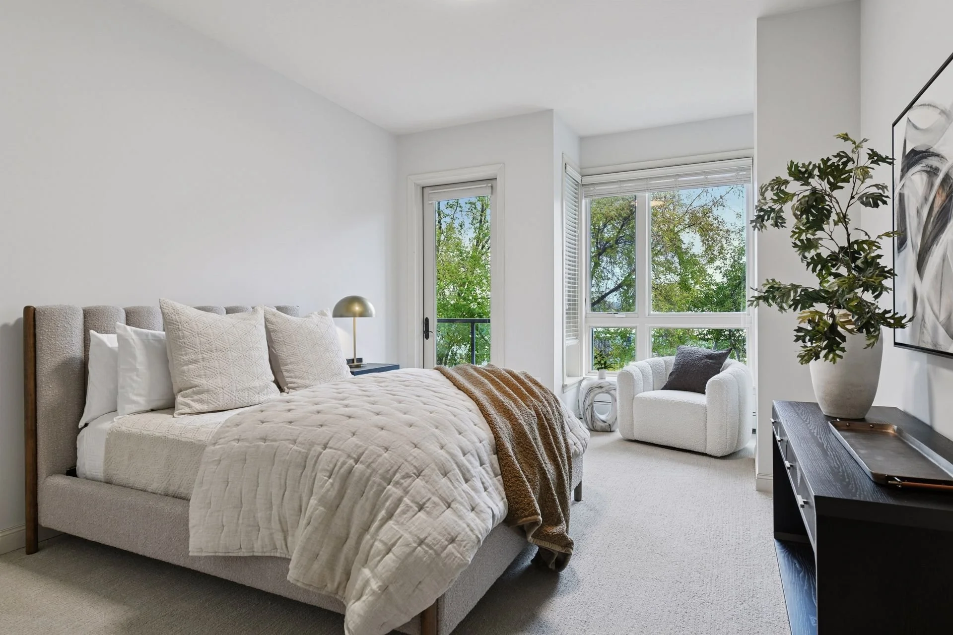 Bright bedroom with a balcony door and large windows revealing green trees outside.