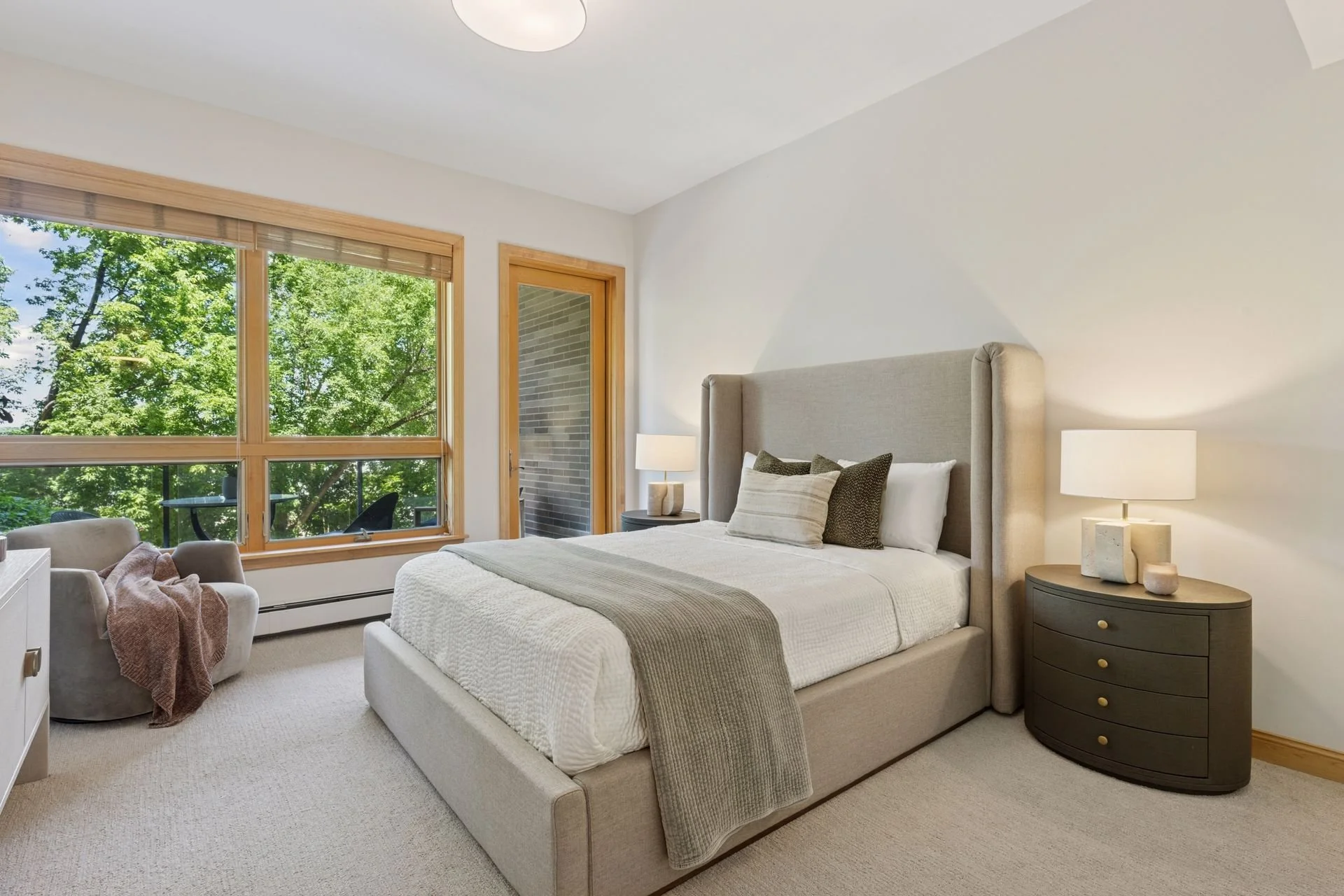 A spacious bedroom. To the left, there's a window with wooden trim showing green trees outside.
