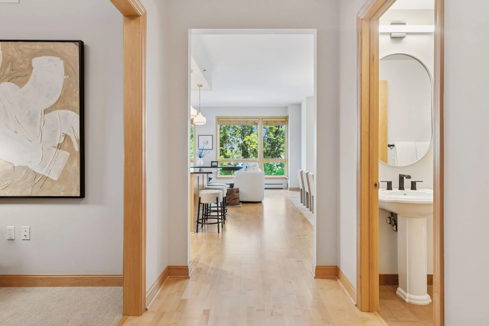 A view of a hallway leading to a living room with large windows. To the right, part of a bathroom with a mirror and wood accents is visible. The walls are white, and the floor is light wood.
