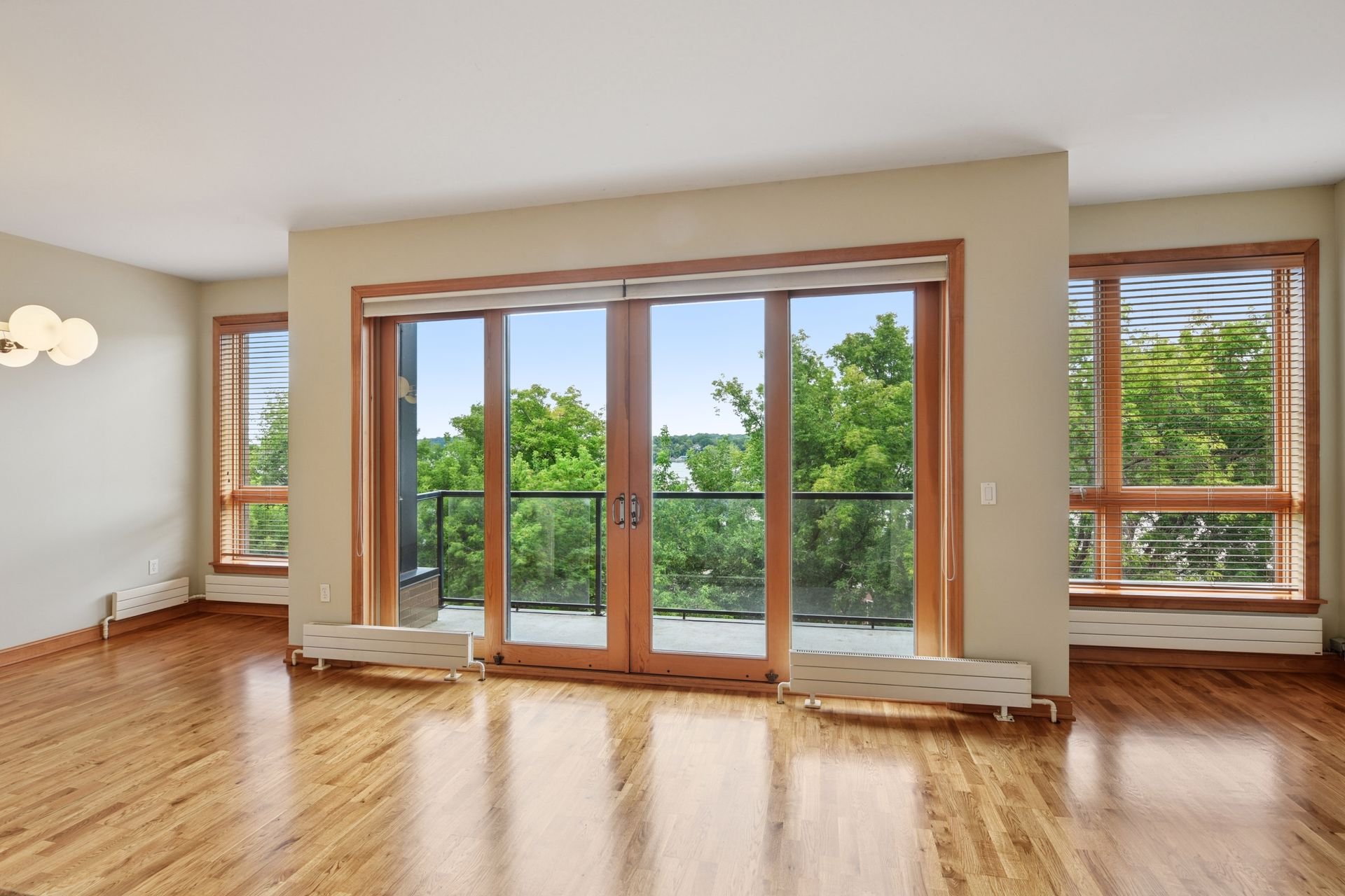 Empty living room with hardwood flooring, large sliding glass doors leading to a balcony, and multiple windows with wooden blinds showing green trees outside.