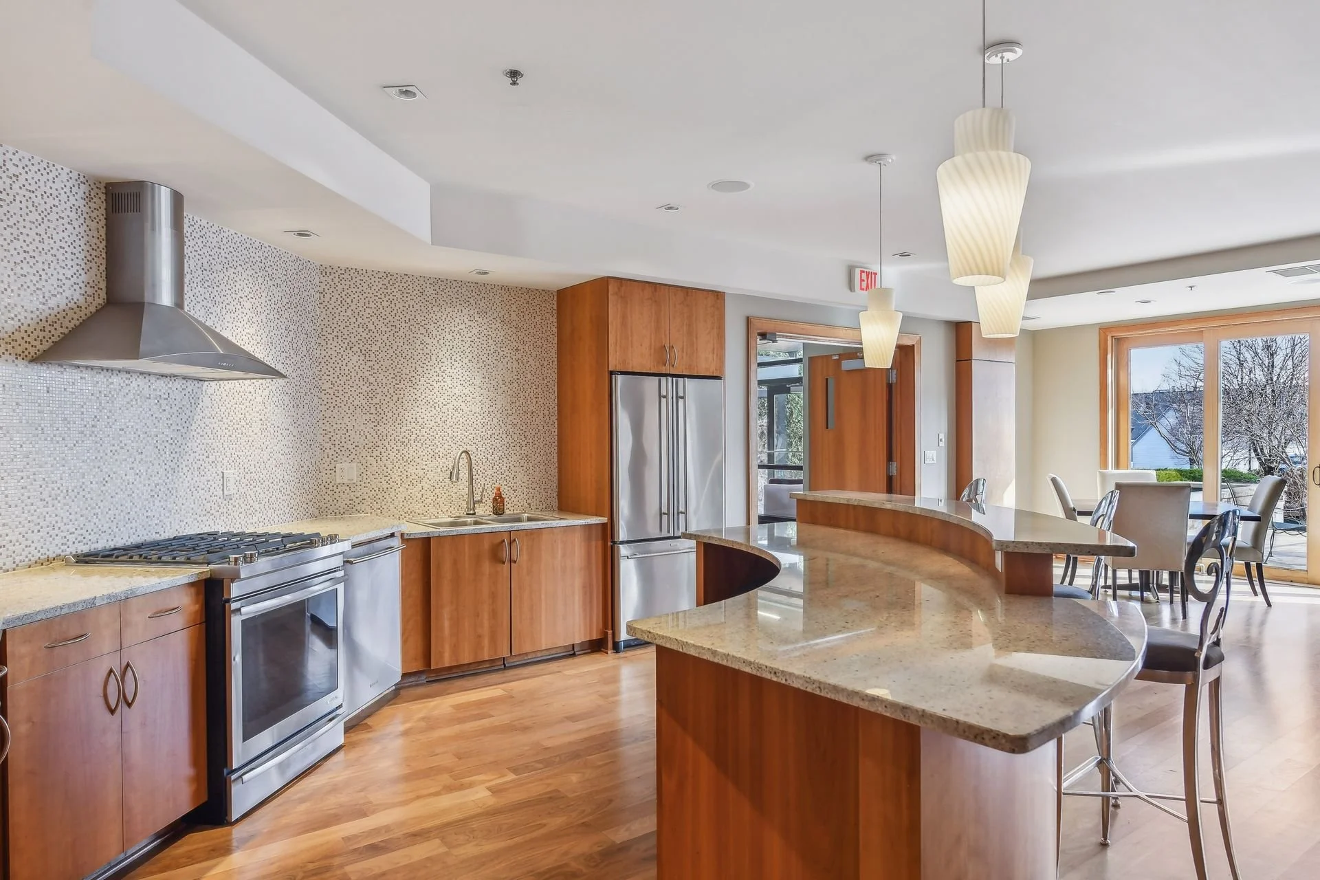 Community room's modern kitchen with wooden cabinets, stainless steel appliances, granite countertops, hardwood flooring, pendant lighting, and large windows with an outdoor view.