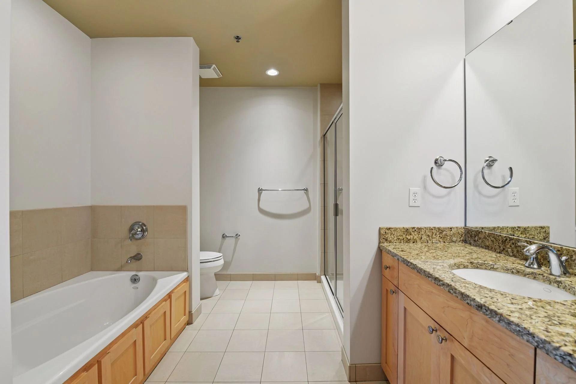 A bathroom with a bathtub, toilet, glass shower enclosure, granite countertop sink, large mirror, towel racks, and tiled floor and walls.