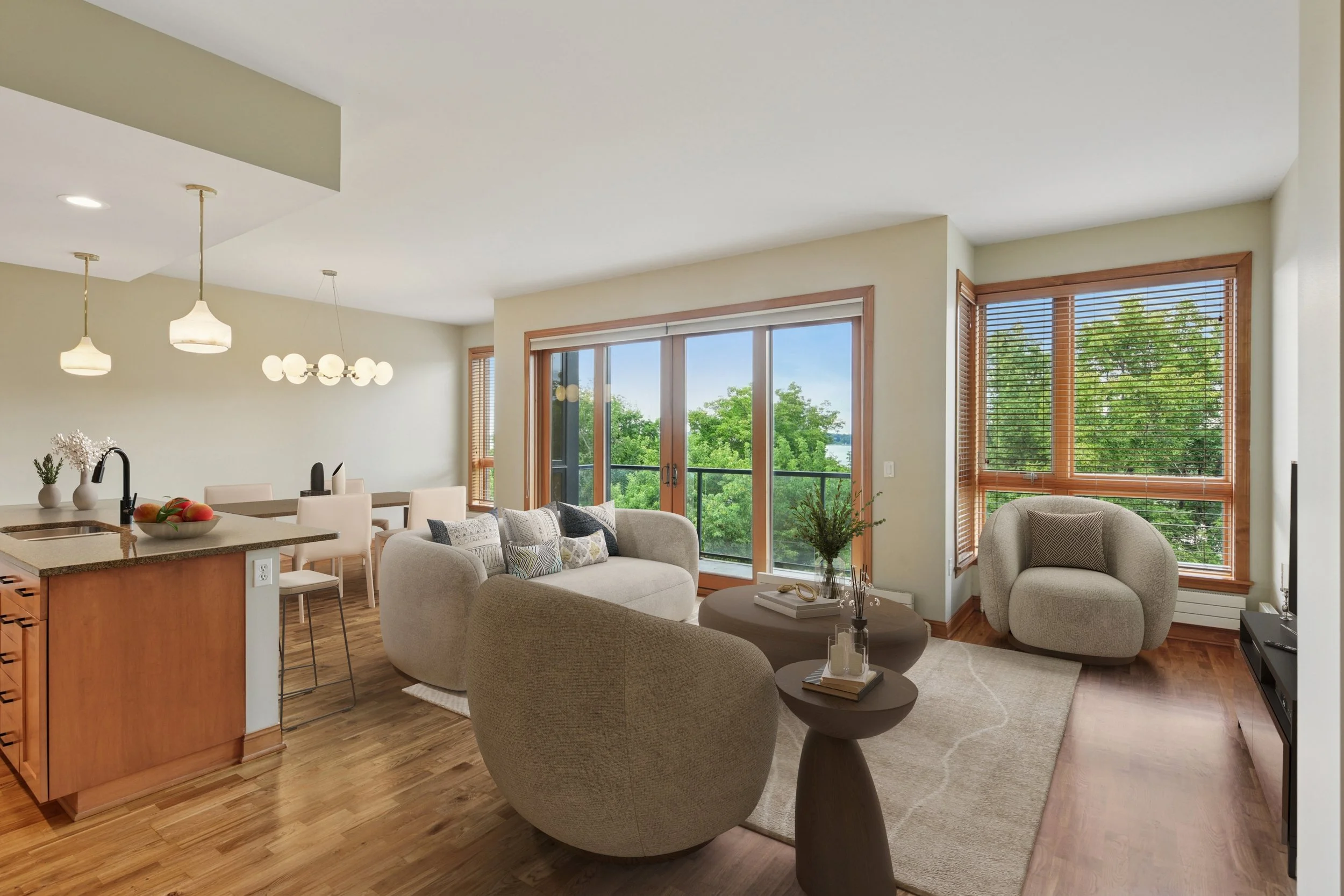 Living room with large windows showing green trees outside. Open kitchen area with a spacious island and hanging lights.
