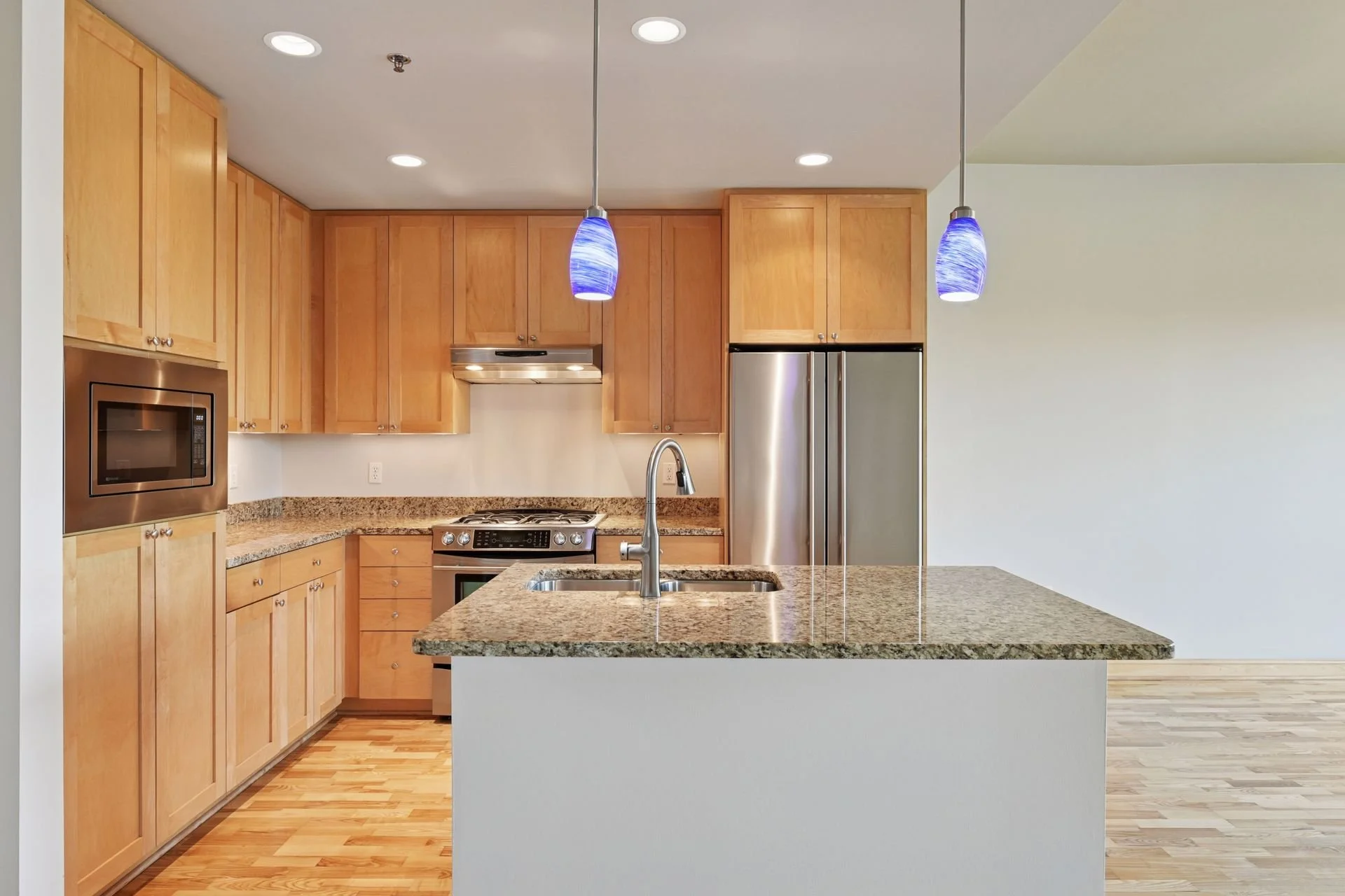 Modern kitchen with wooden cabinets, granite countertops, stainless steel appliances, and two blue pendant lights over the island.