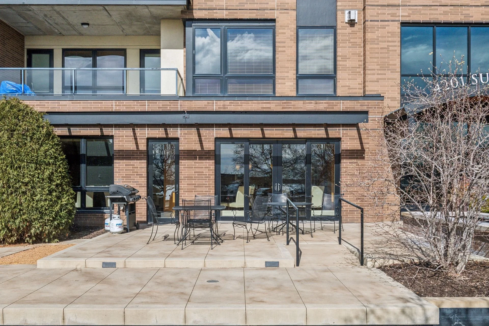 A brick building with glass doors and windows, an outdoor patio with black metal tables and chairs, a barbecue grill, and a bare tree on the right side.