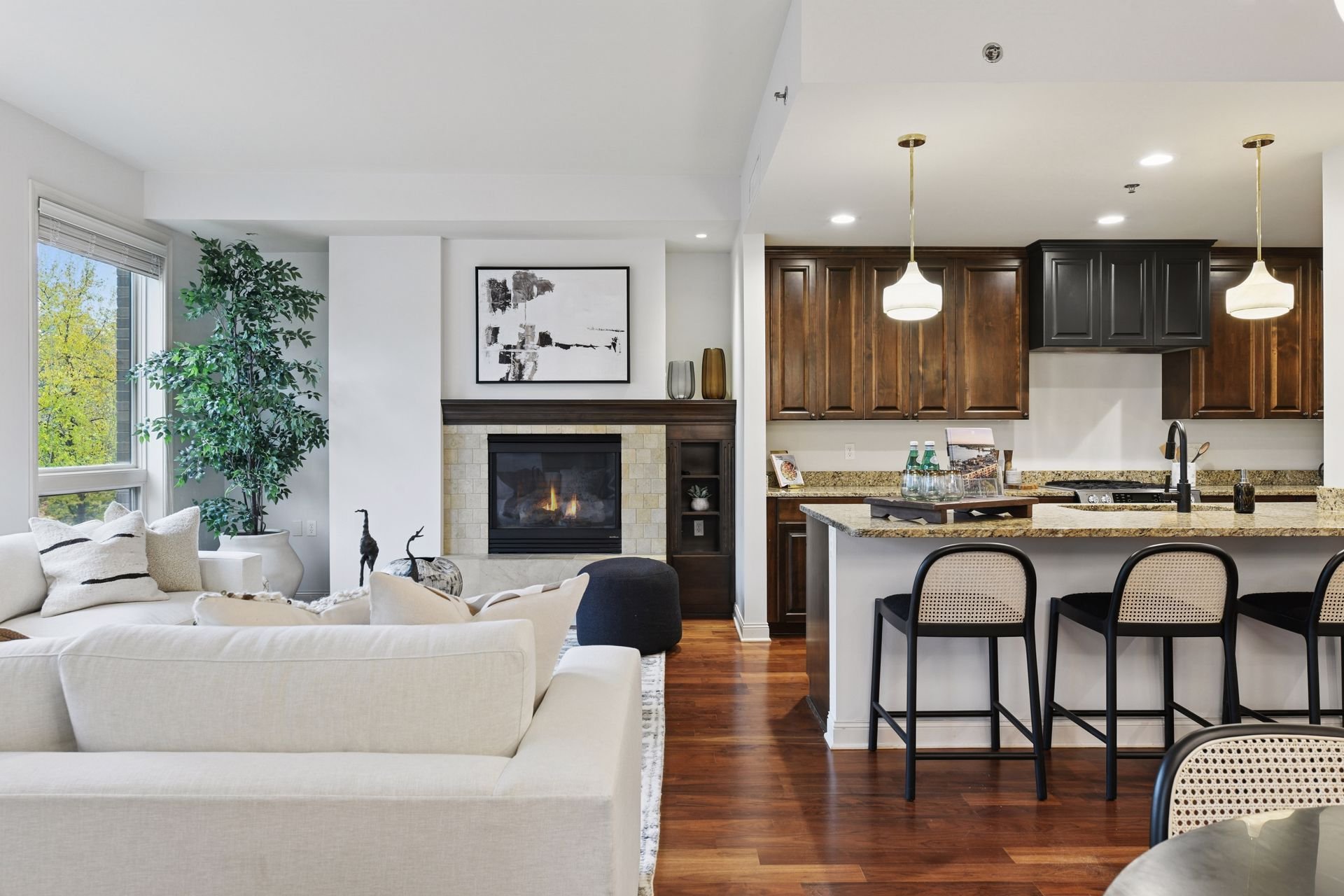 Living room with fireplace, large windows, kitchen with wooden cabinets, granite countertops, and pendant lights.