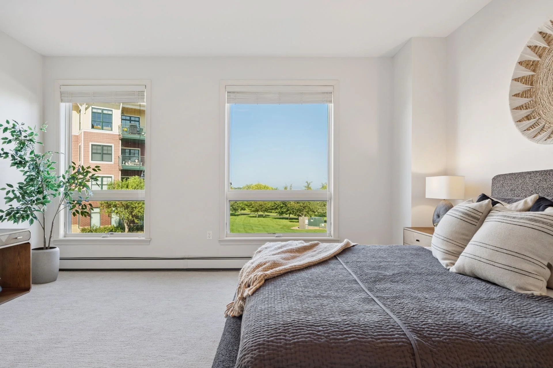 A bright, minimalist bedroom with two large windows showing a green park and a modern apartment building outside.