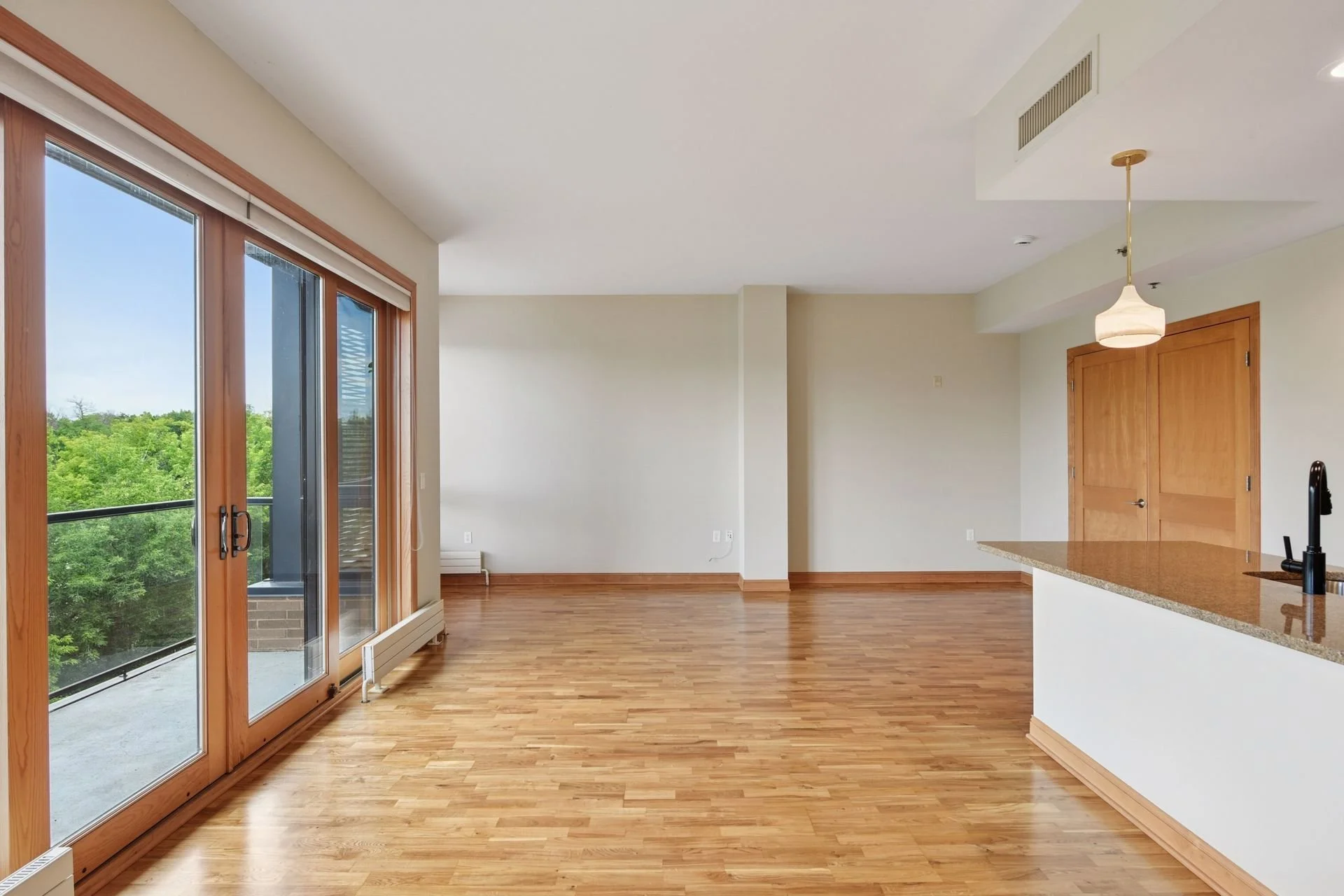 Empty living room and kitchen with hardwood floors, large sliding glass door leading to balcony with trees outside, white walls, wooden accents, and a hanging ceiling lamp.