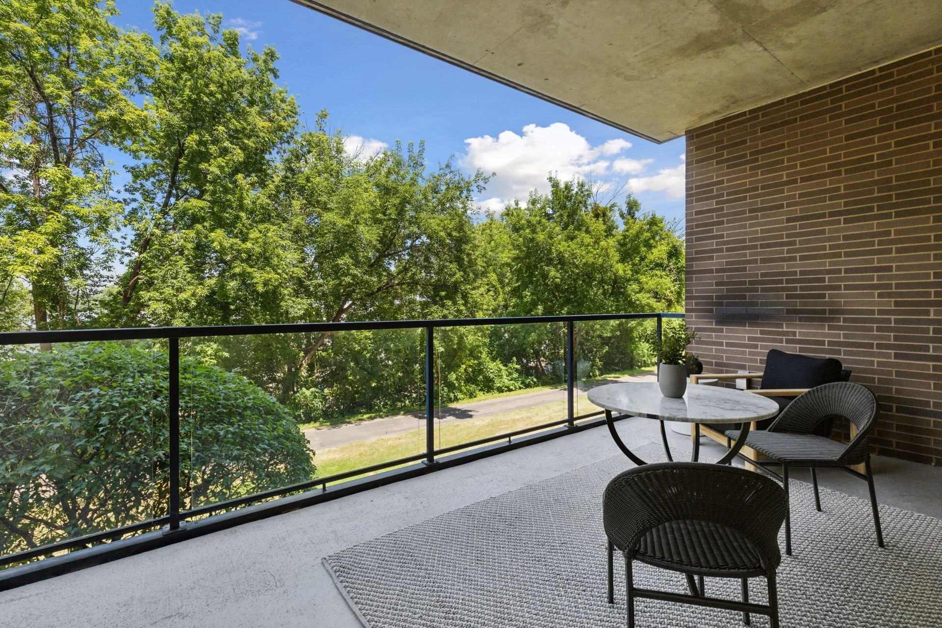 Balcony with black metal railing, against a brick wall, overlooking a lush green tree and blue sky.