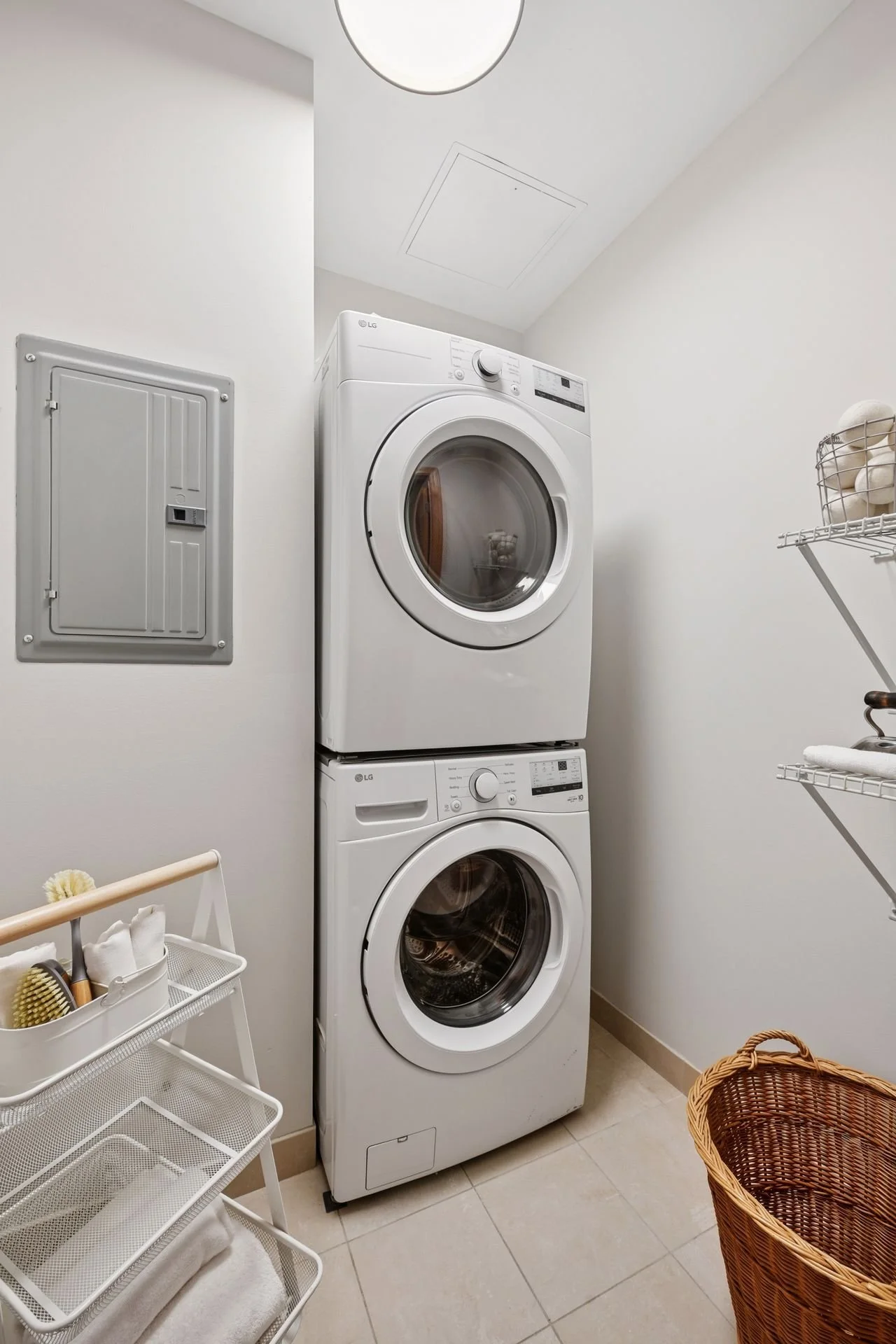 A laundry room with a stacked washer and dryer, a wire shelf, and a utility box on the wall.