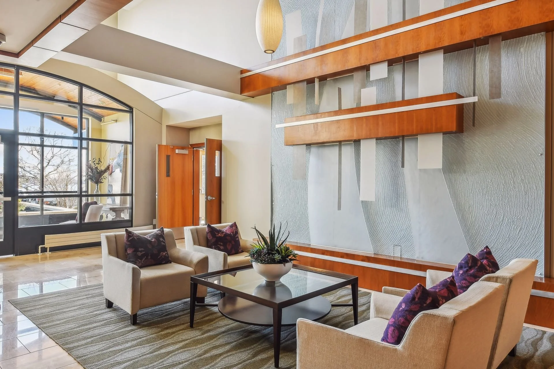 Lobby with beige sofas, purple pillows, a glass coffee table with a plant, large front window, wooden walls, and a decorative wall art piece.