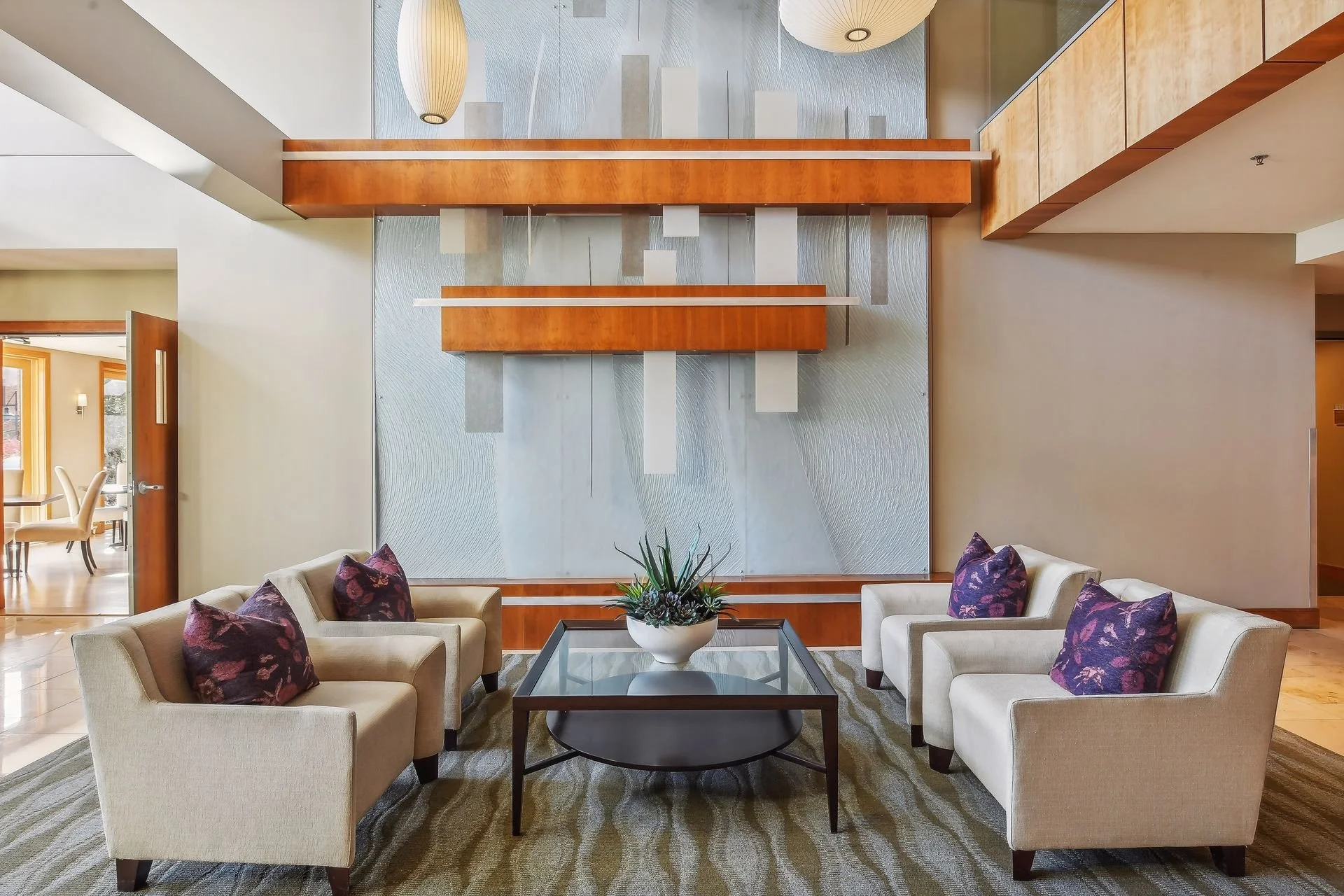Lobby area with four cream-colored sofas, each with purple patterned pillows, arranged around a glass-top coffee table with a white ceramic pot holding a green plant, against a textured blue-gray accent wall with wooden and metal decorative panels.