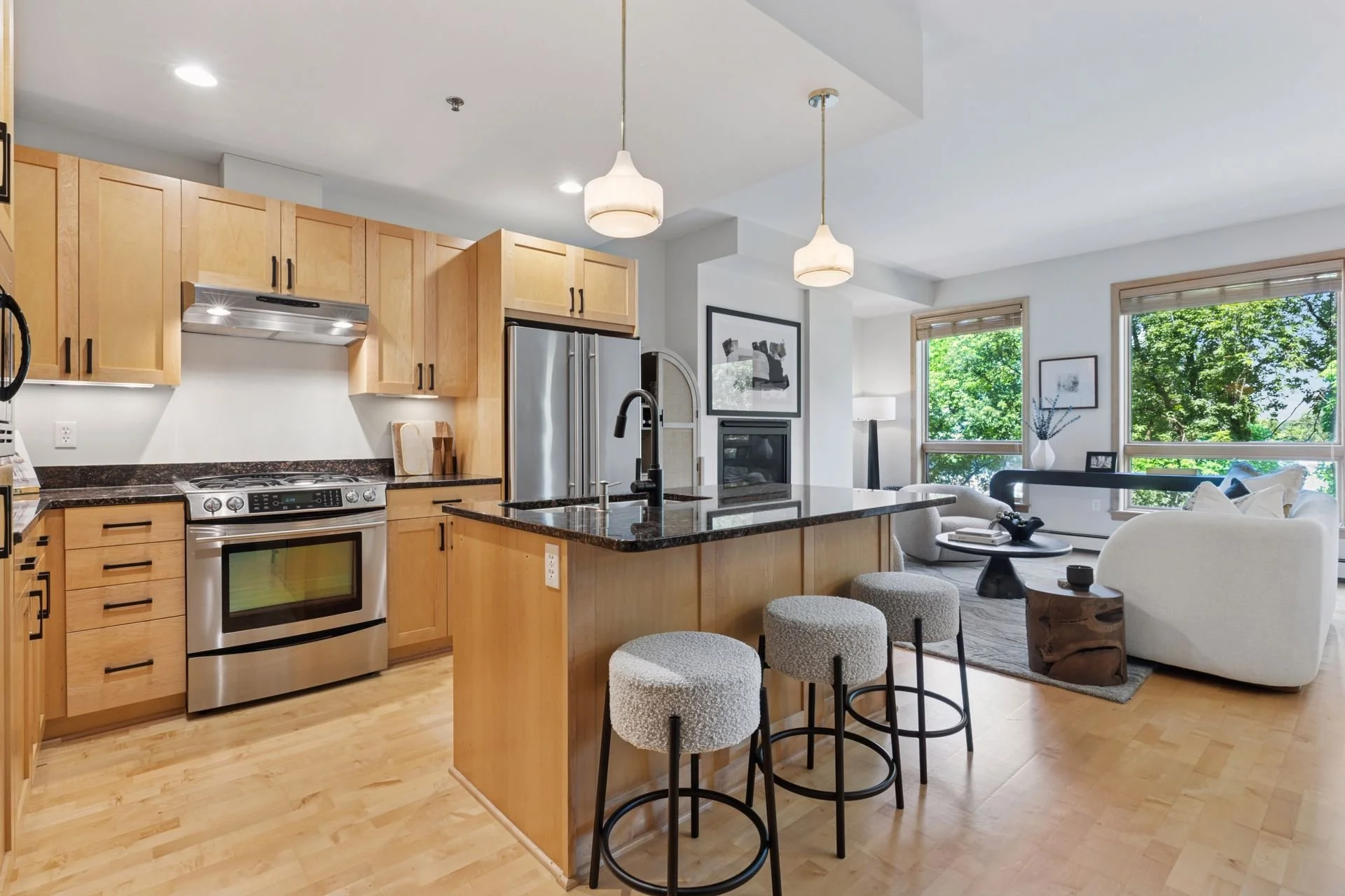 Open-concept kitchen with light wood cabinets, black granite countertops, stainless steel appliances, and a spacious island. Adjacent living room with large windows.