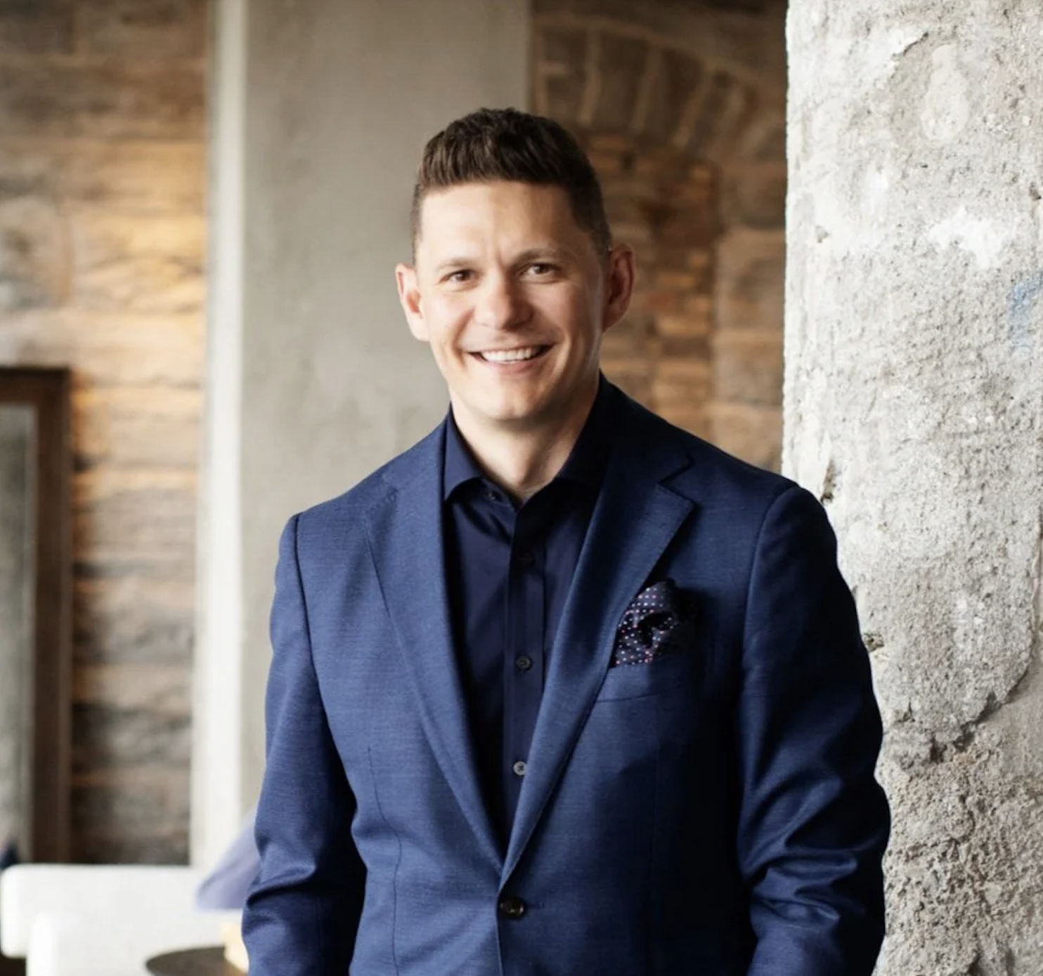 A smiling man in a navy blue suit and matching shirt, standing against a stone wall in an indoor setting.