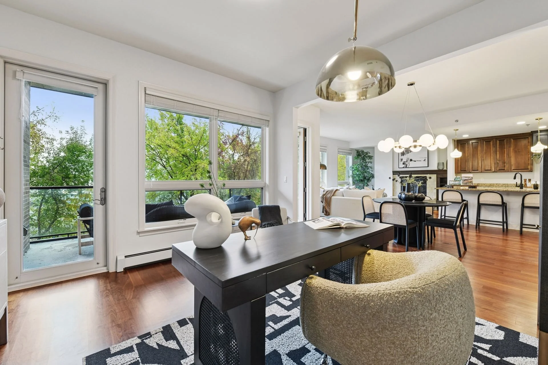 An open concept living and dining area with large windows showing green trees outside, hardwood floors, and contemporary lighting fixtures.