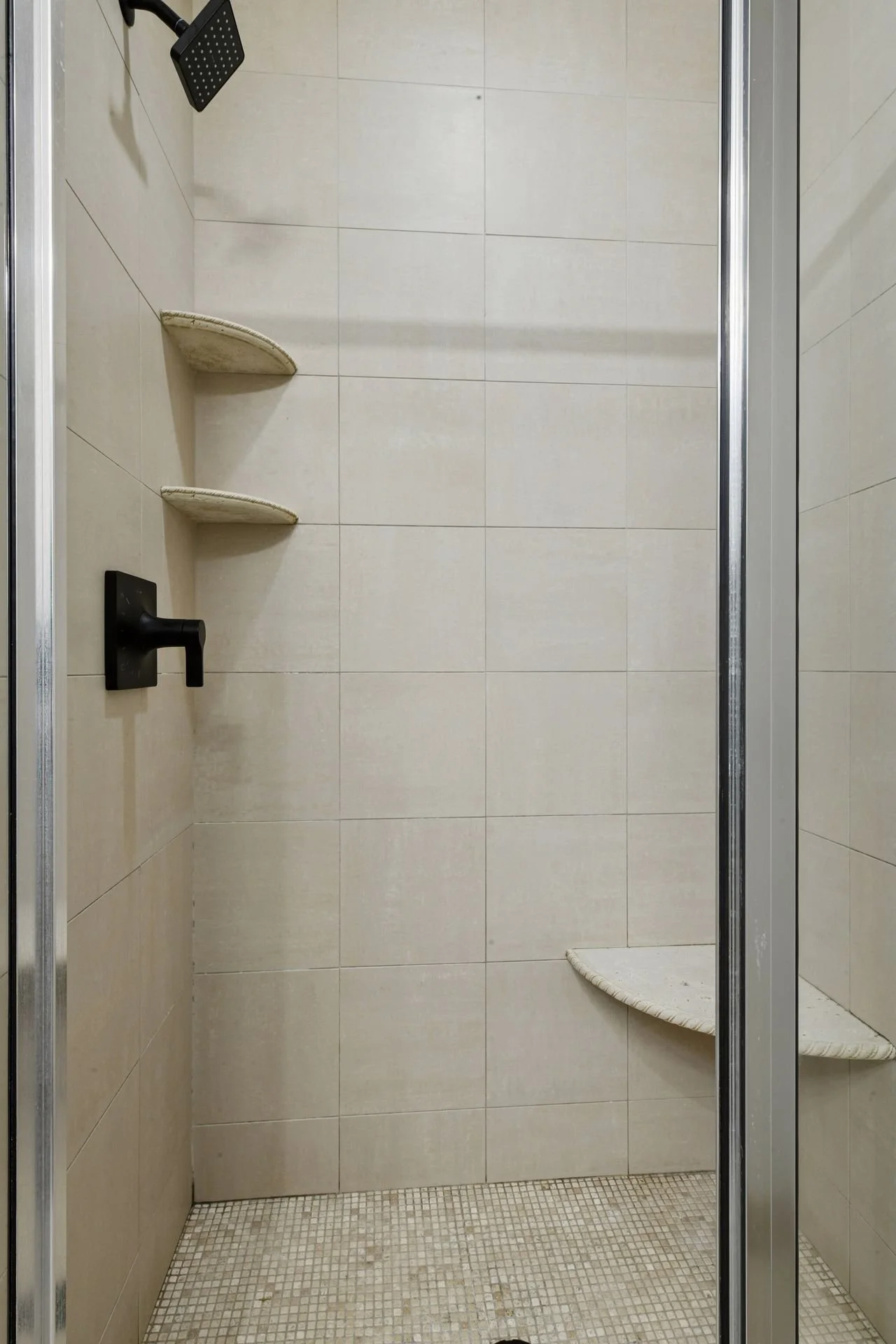 Empty shower with beige tiled walls, built-in corner shelves, a black showerhead, a black faucet, and a glass door with a metal frame.