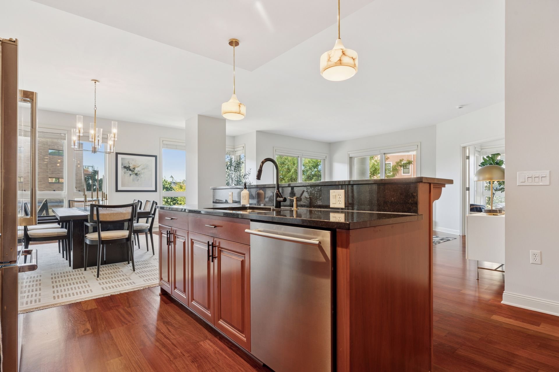 Open-concept kitchen with wooden cabinets, black granite countertops, stainless steel dishwasher, and a view of a dining area with pendant lighting, and large windows letting in natural light.
