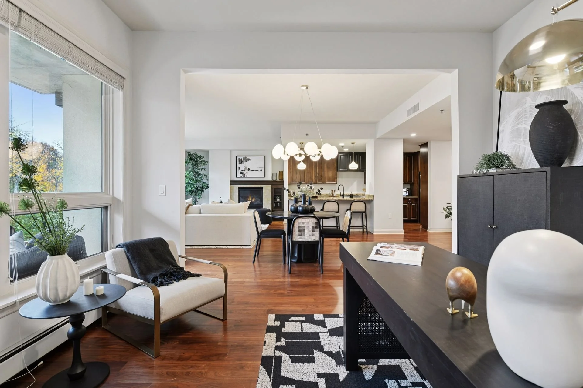 Interior of a modern living room and dining area with a view into the kitchen, featuring wooden floors, white walls, large windows, and a hanging light fixture.