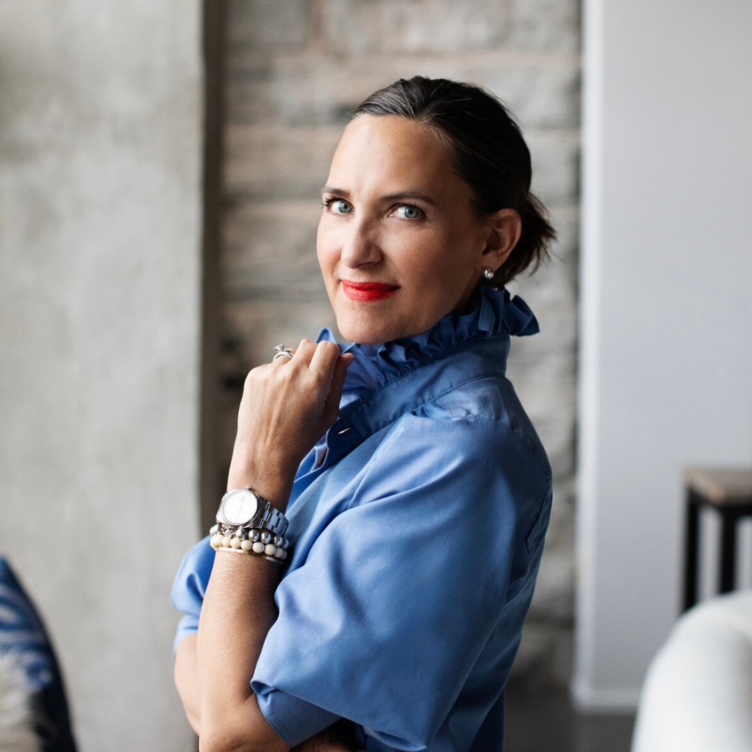 A woman with dark hair, blue eyes, and red lipstick, wearing a blue blouse with ruffled collar, sitting indoors with a background of exposed brick wall and white wall, looking at the camera.