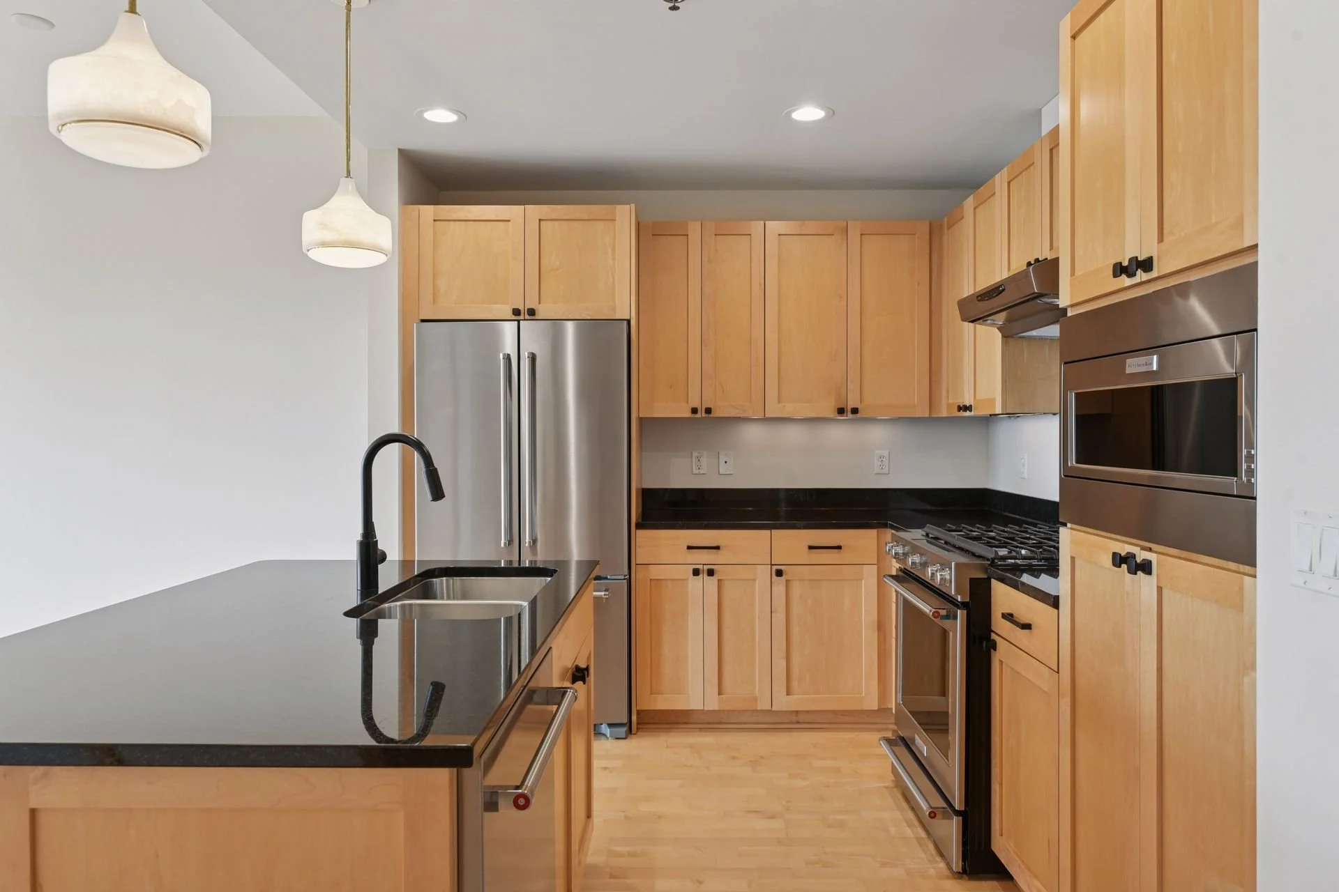 Modern kitchen with wooden cabinets, black countertops, stainless steel refrigerator, oven, microwave, and black sink with faucet.