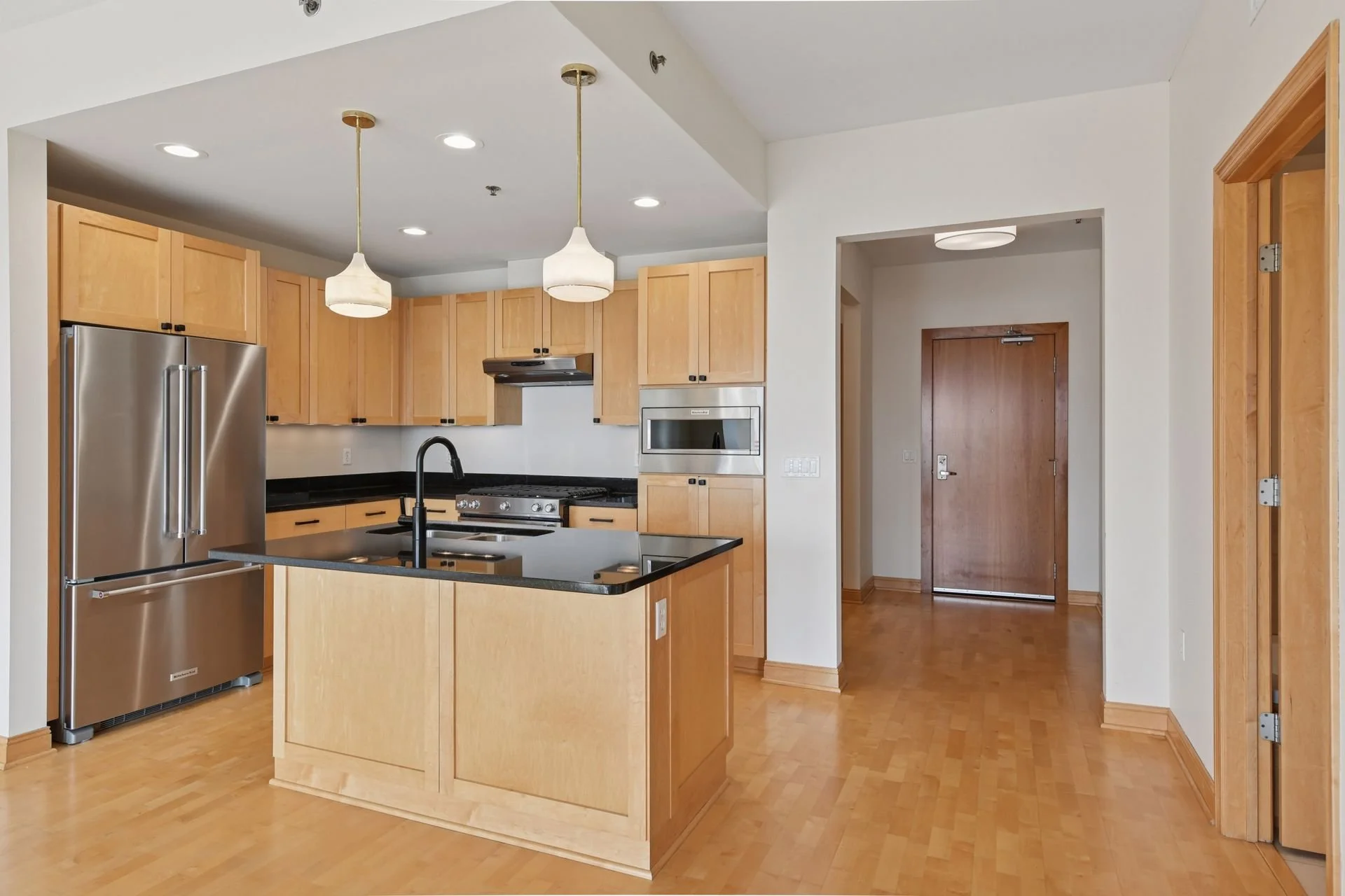 Modern kitchen with light wood cabinets, black countertops, stainless steel refrigerator, and a kitchen island with a black sink and faucet, illuminated by pendant and recessed lighting.