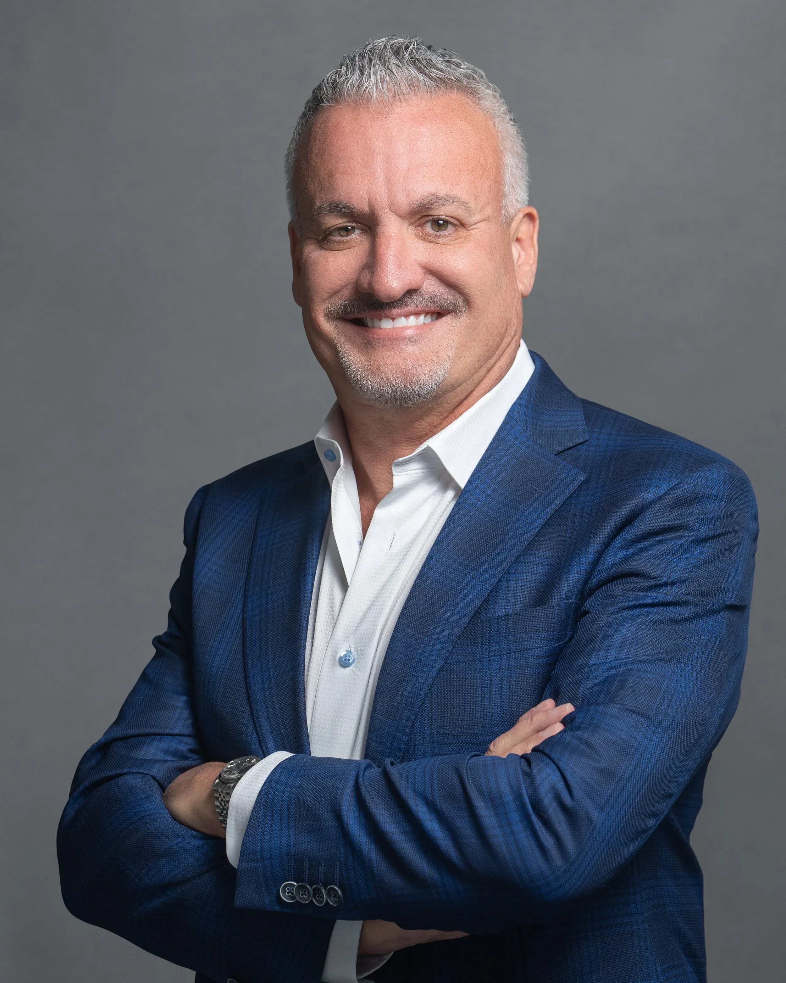A middle-aged man with short gray hair and a trimmed goatee smiling confidently. He is wearing a blue suit jacket over a white dress shirt and has his arms crossed against a plain gray studio background.