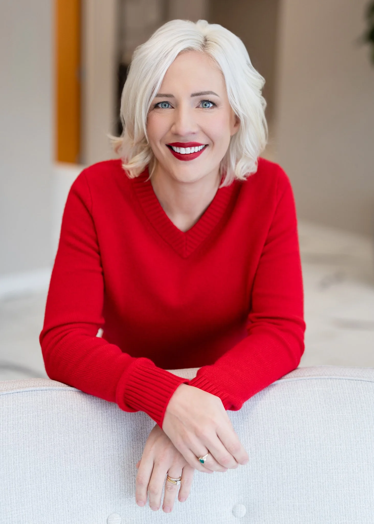 A woman with blonde hair and blue eyes smiling, wearing a red sweater, sitting with her hands resting on a light-colored couch.