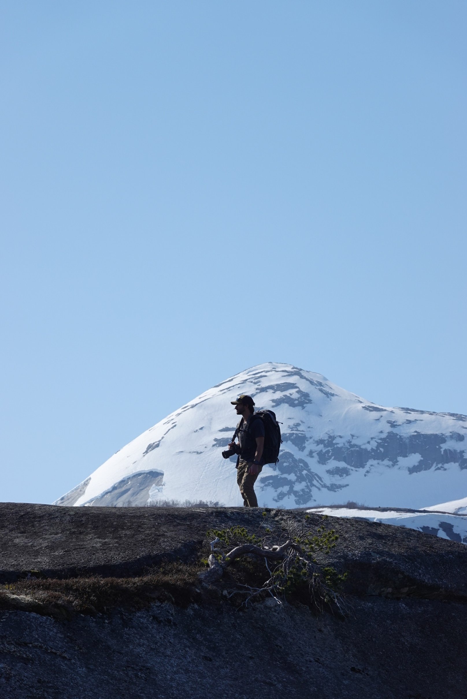 A hiker with a backpack and camera walking on a rocky terrain with a snow-capped mountain and clear blue sky in the background.