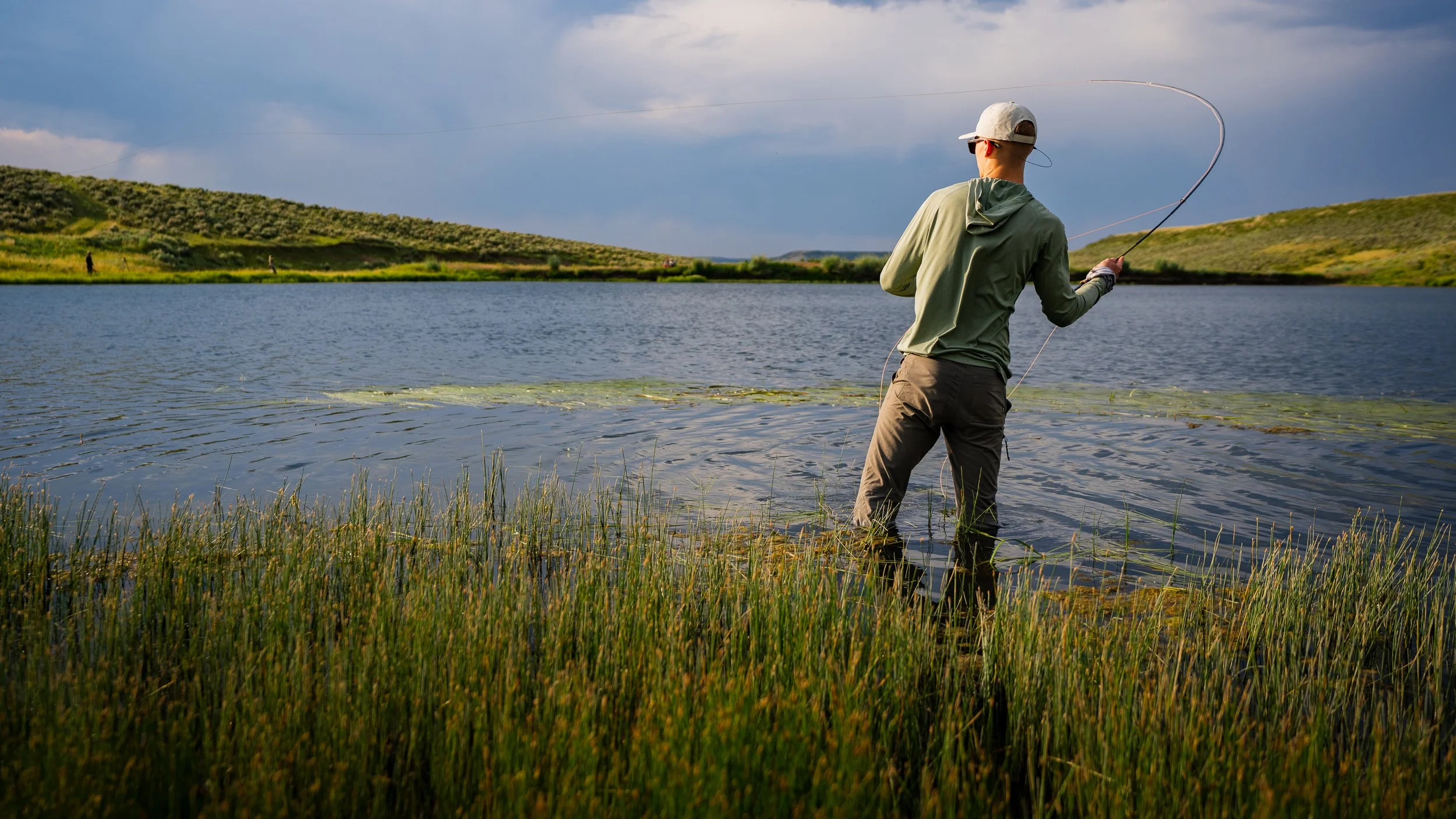 Man fishing in a lake with grassy shoreline and hills in the background during daytime.