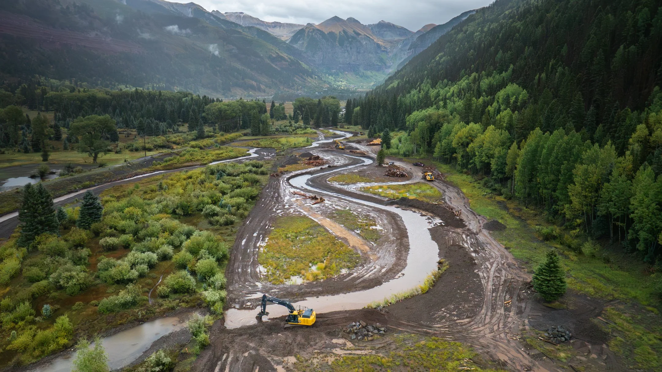 A drone image of a Trout Unlimited Construction site restoring a river and willows/stream vegetation in a lush green valley with a river (Telluride Valley), surrounded by dense forested mountains,  with excavators working.