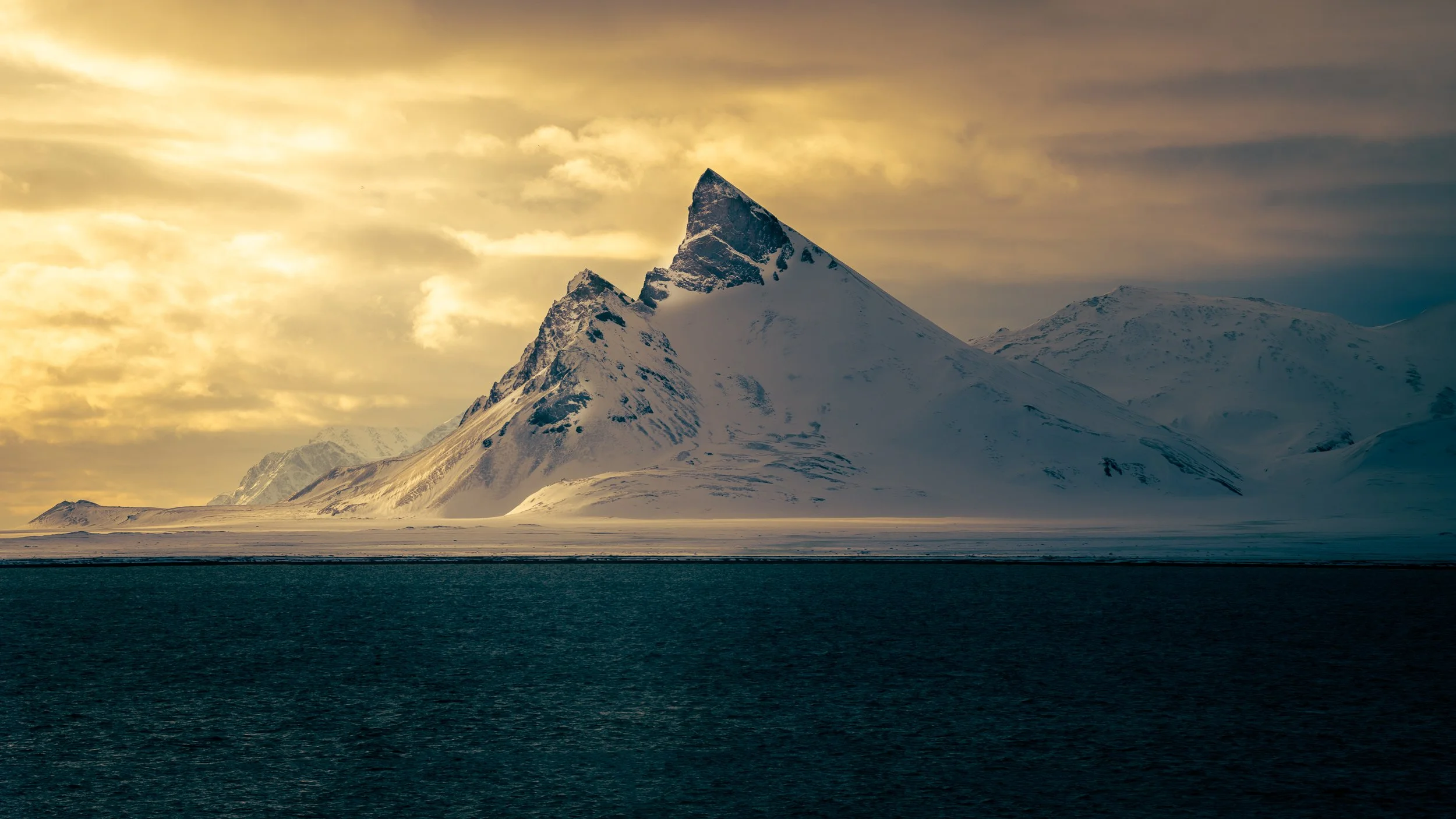 Snow-covered mountain peaks with a triangular-shaped summit, overlooking a dark body of water under a cloudy sky with a warm light.