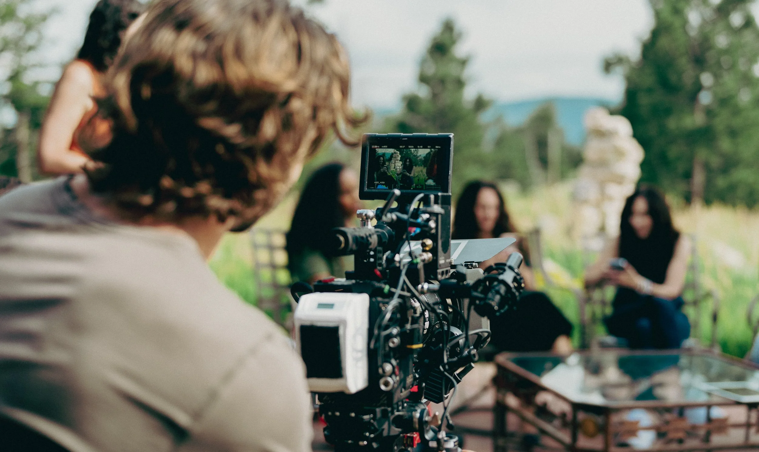 A person operates a camera filming three women sitting outdoors on benches, with trees and a sculpture in the background.